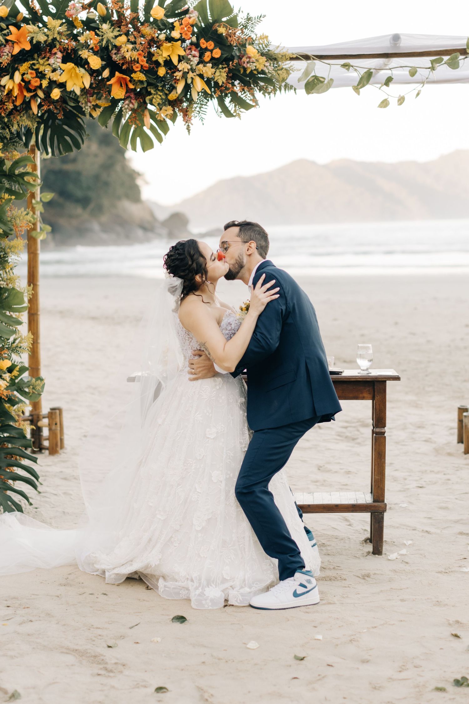 Intimate wedding moment captured under a lush floral arch on a secluded beach at sunset.