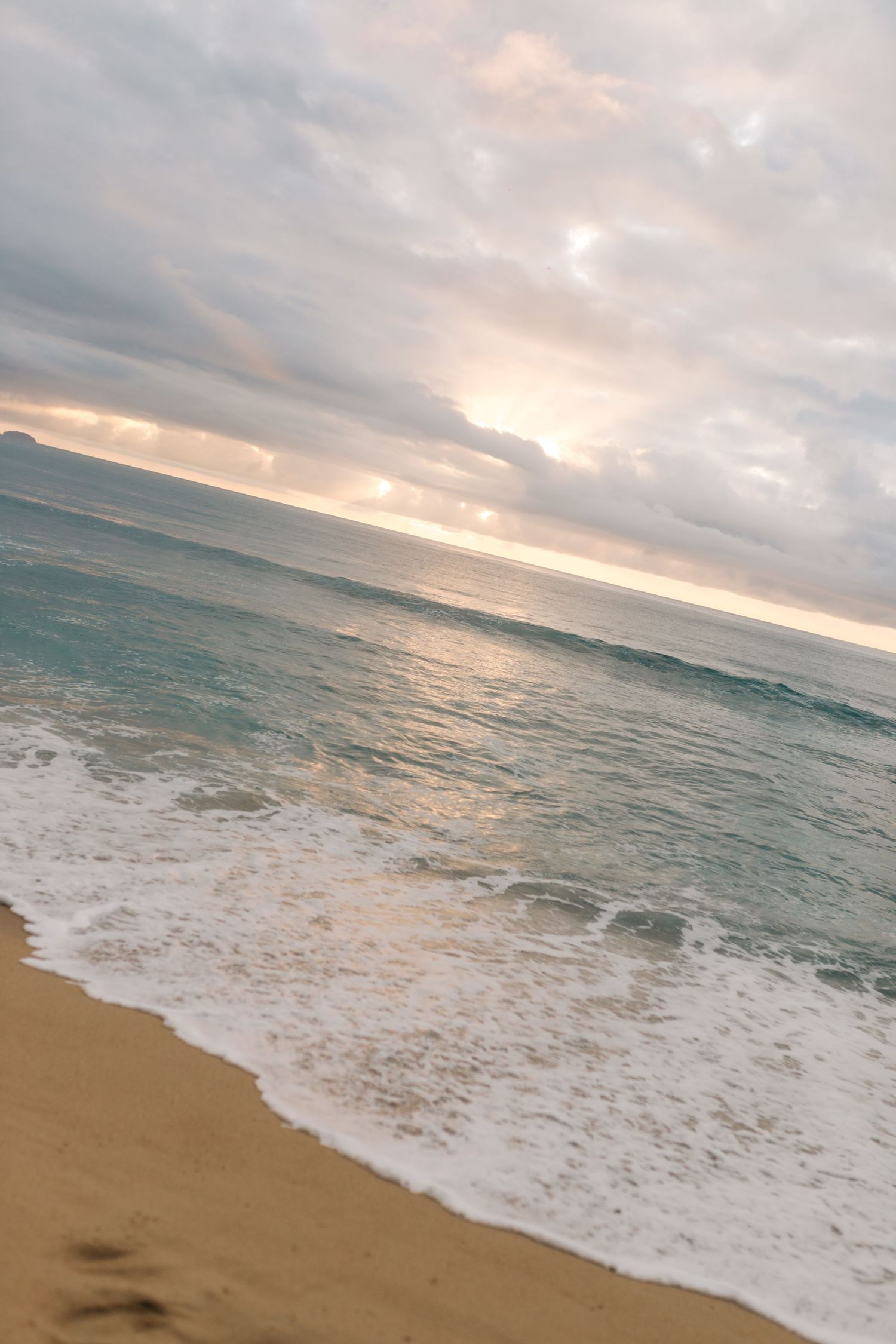 Waves crash onto a golden sandy beach during sunset with cloudy skies.