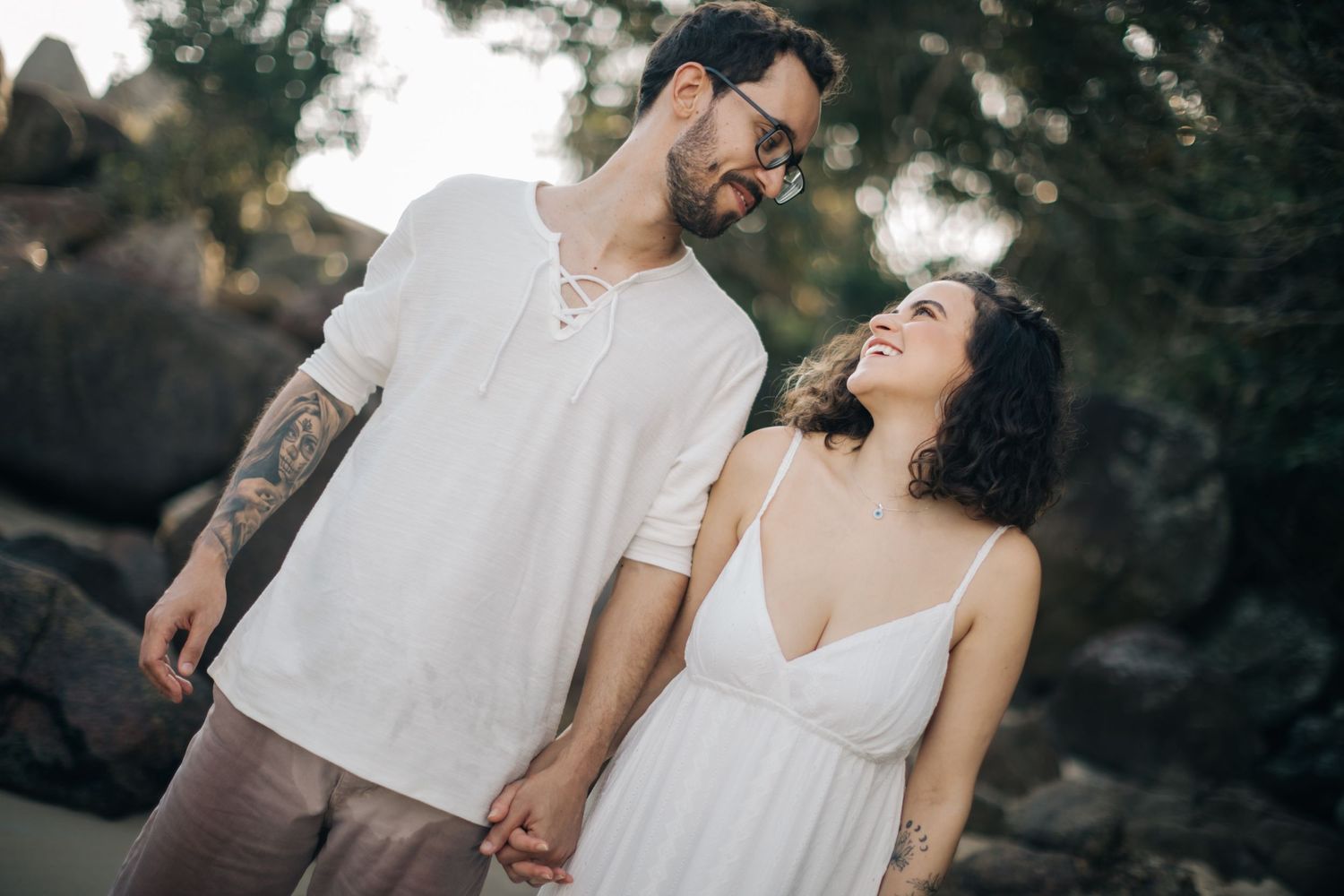Couple laughing together on beach with rocky backdrop in coordinated white outfits.