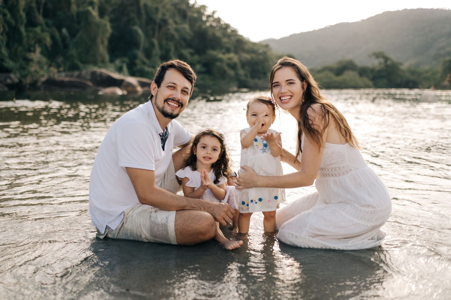 A family of four dressed in white sits together at the water's edge during sunset.