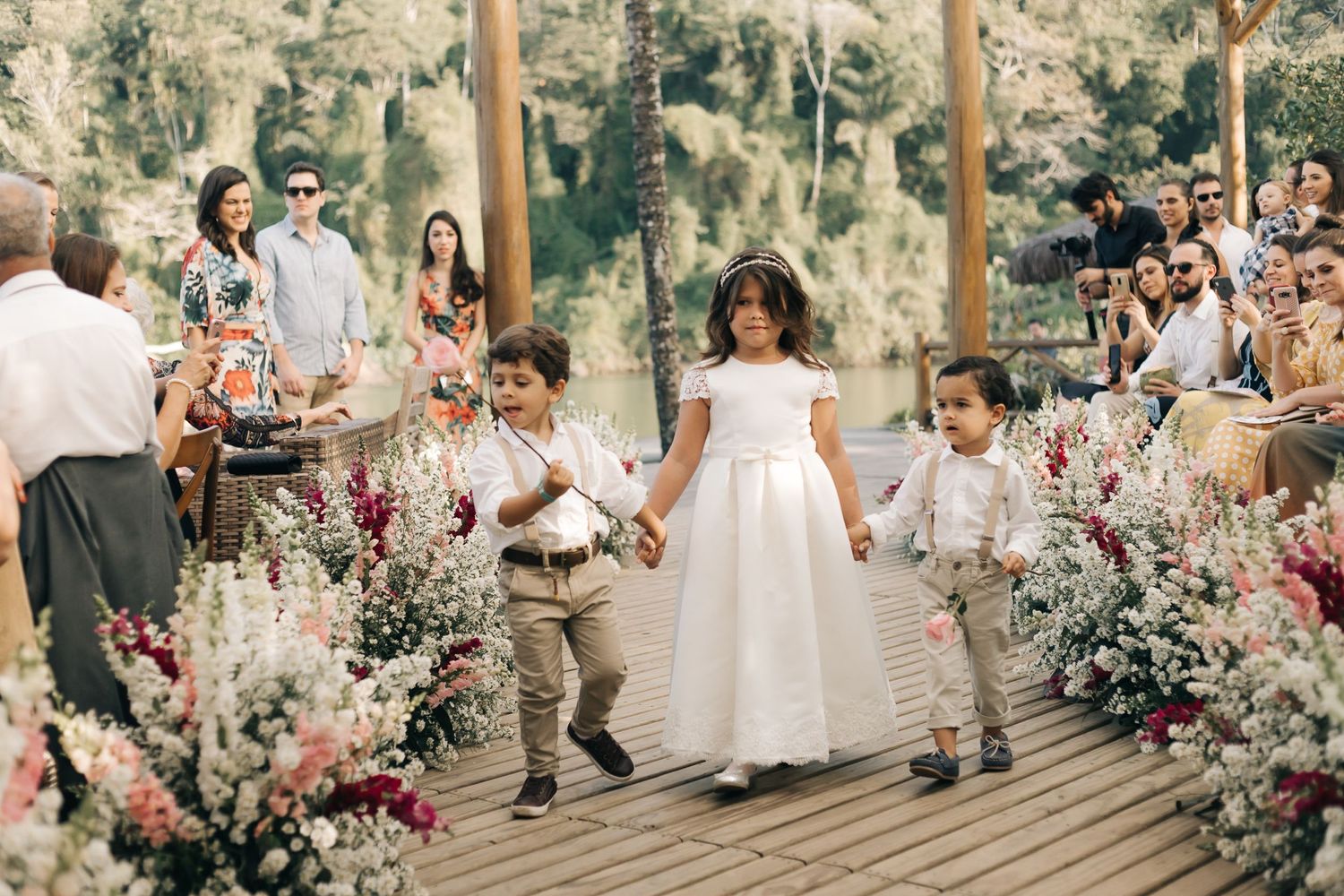 Children walking down floral-lined aisle during wedding ceremony on wooden deck.