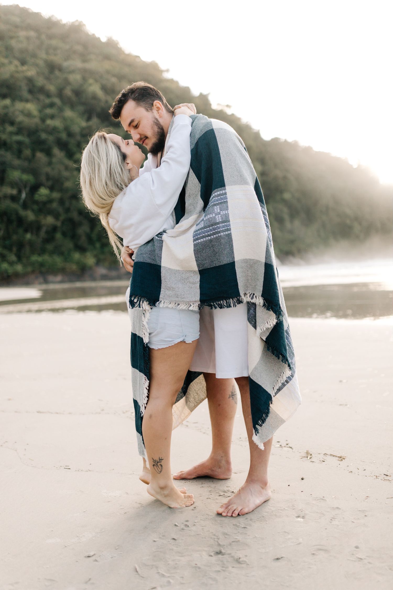 A couple embraces tenderly while wrapped in a navy and white blanket on a foggy beach.