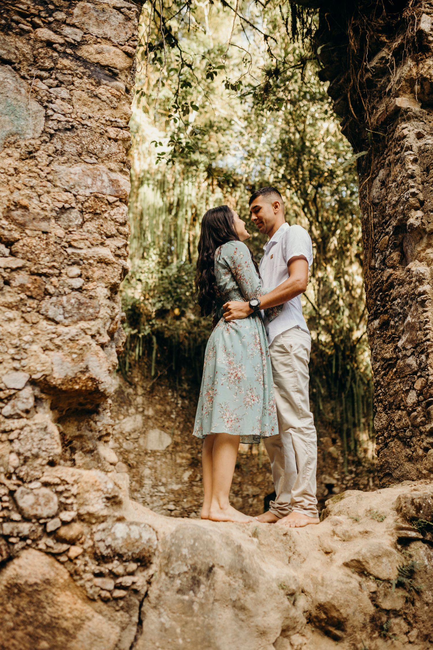 A romantic embrace atop stone ruins with sunlit foliage in the background.