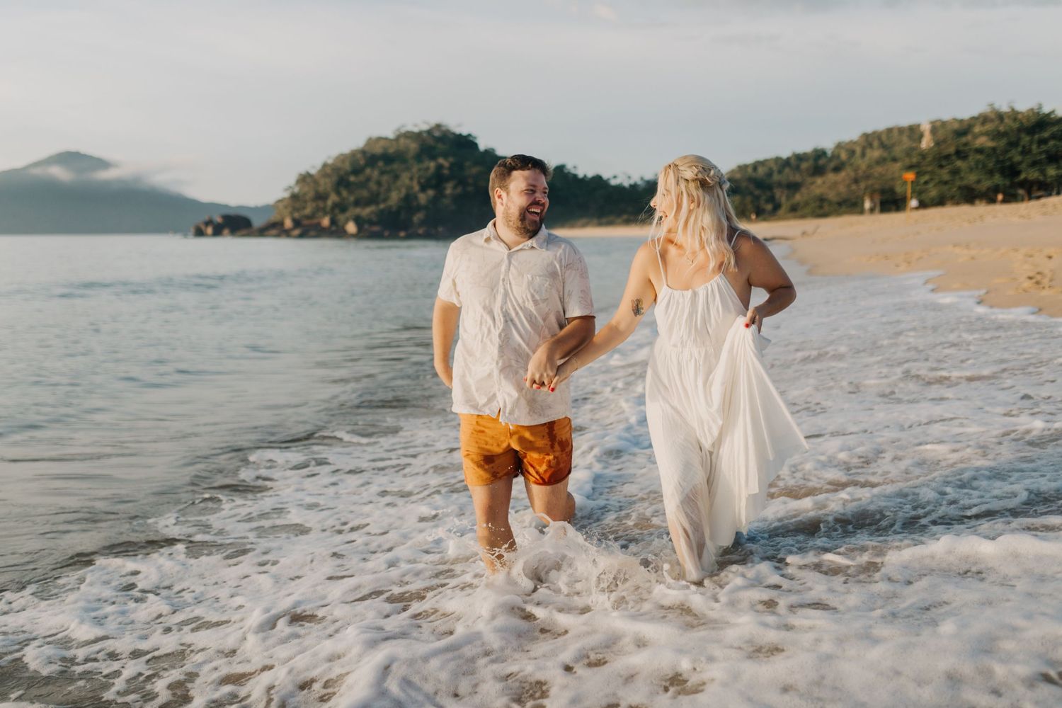 Couple walking along a scenic beach shoreline at sunset, leaving footprints in the wet sand.