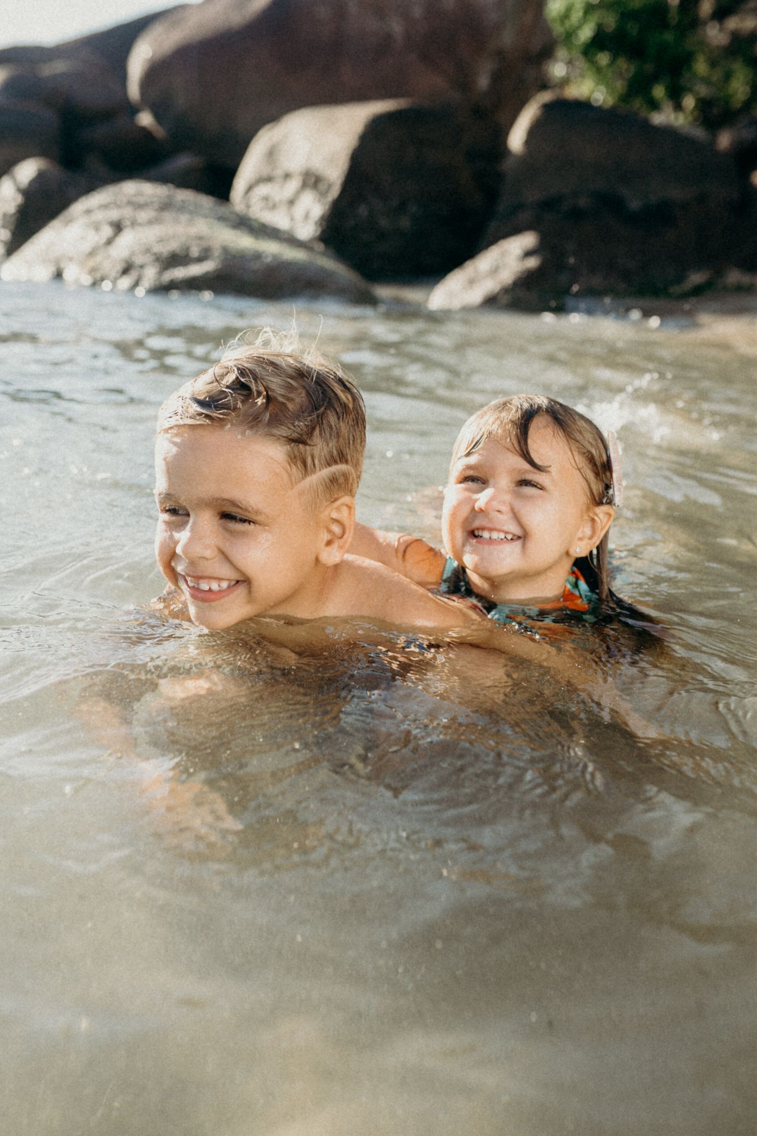 Two children laughing while playing in ocean water near rocky shore.