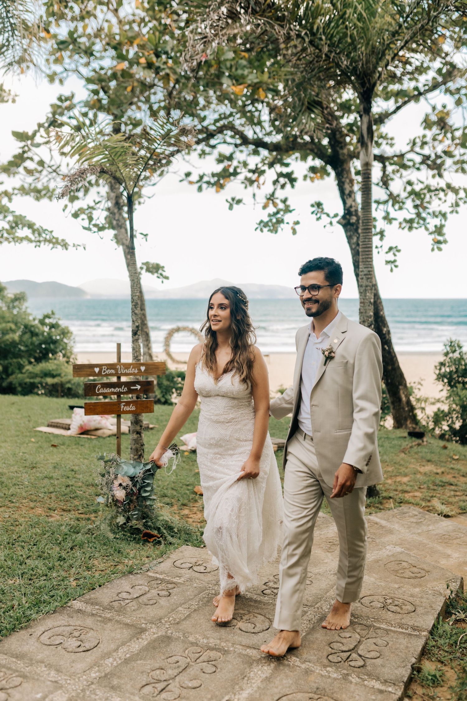 A wedding couple walks along a beachside path with trees and ocean views.