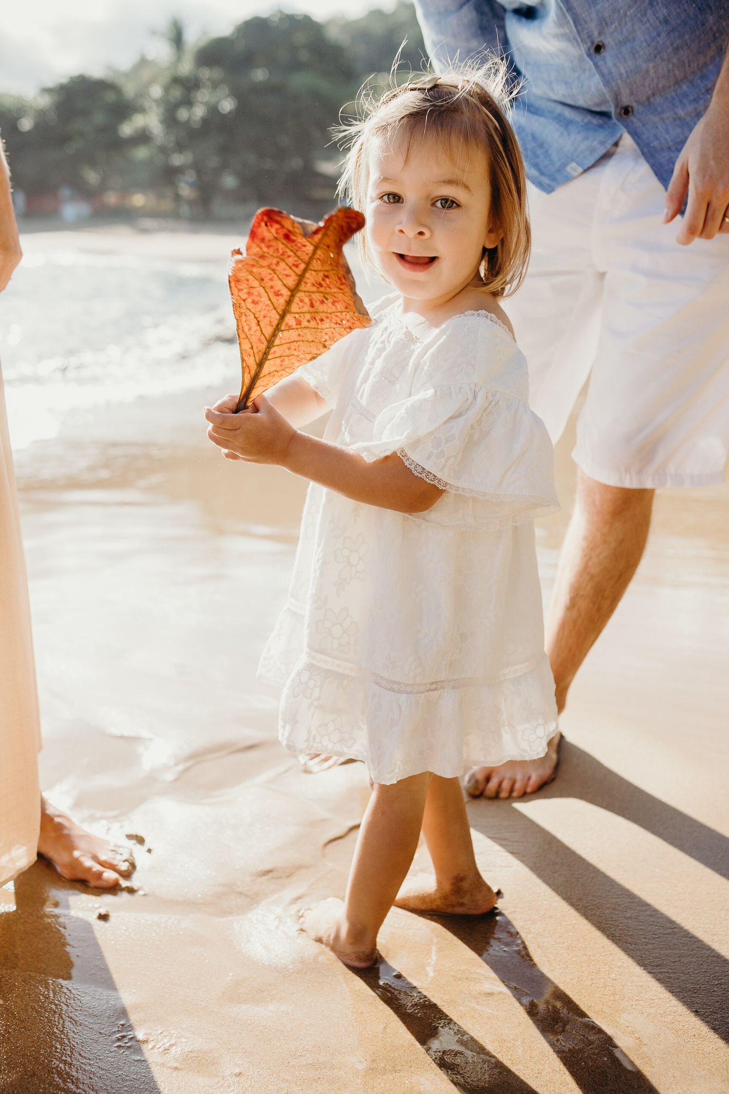 A small child in white dress holds large orange leaf while playing on beach.