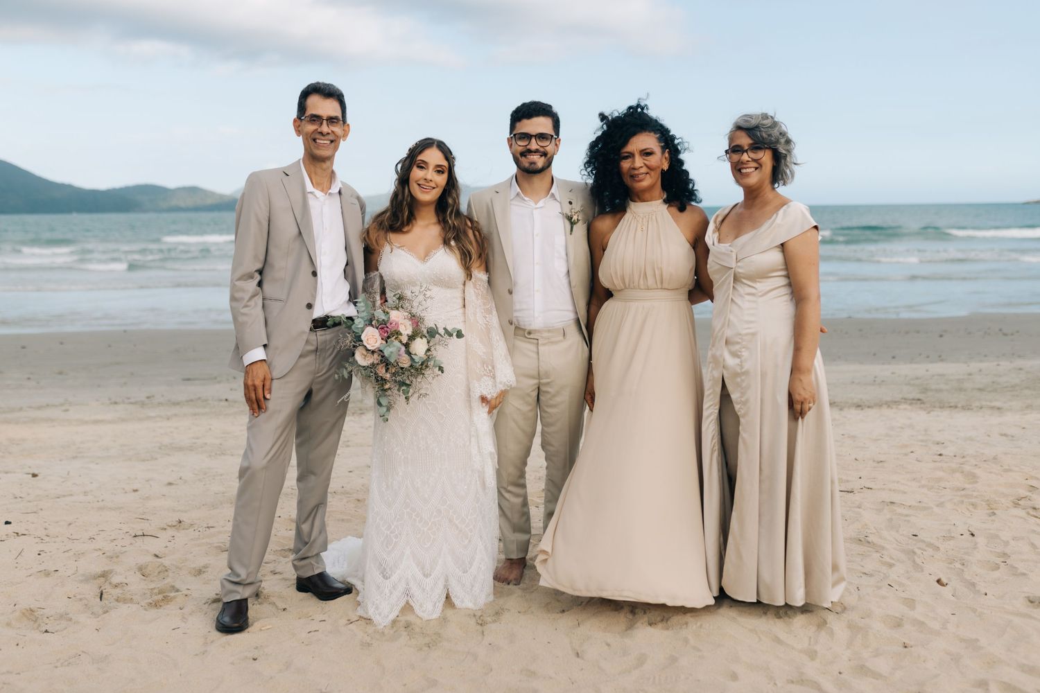 Wedding group poses casually on beach with ocean waves and mountains in background.