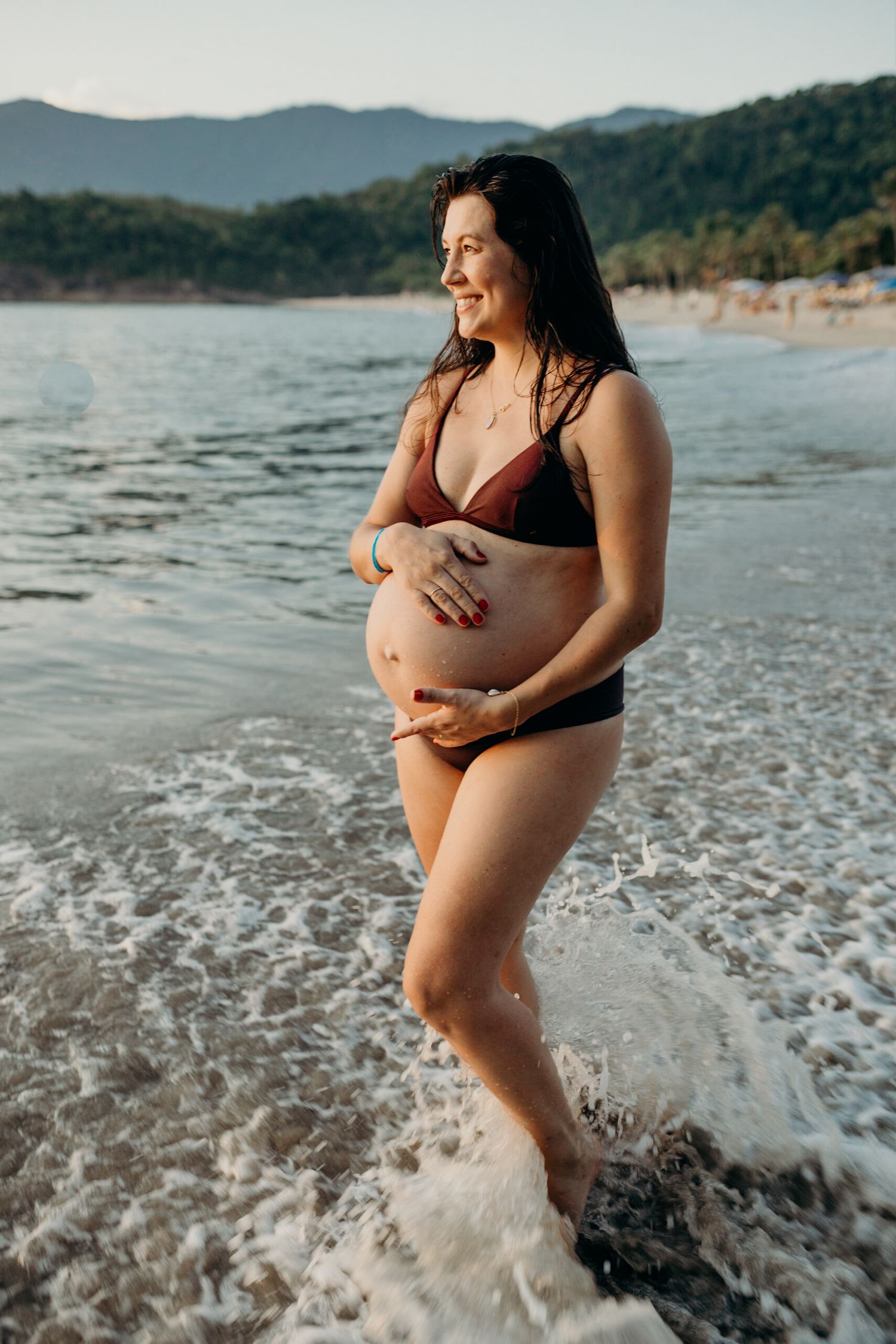 Mother-to-be standing in ocean waves during sunset at beach with mountains in background.