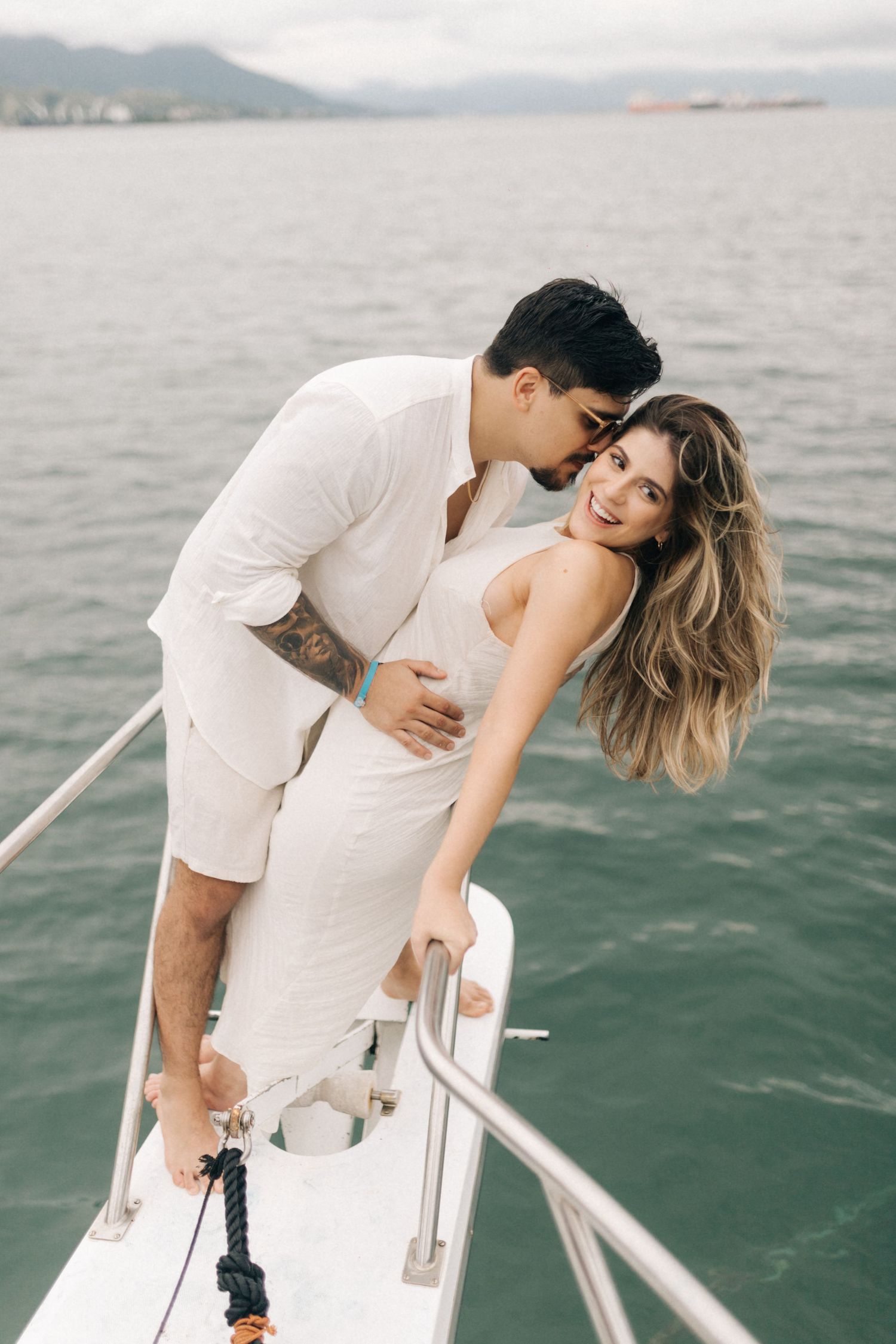 A couple dressed in white embraces on the bow of a boat overlooking calm waters.
