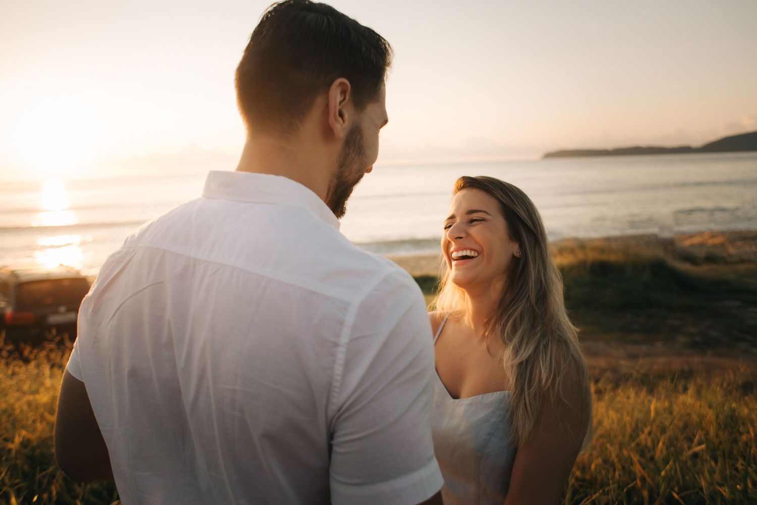 Joyful couple laughing together against beautiful coastal sunset backdrop.