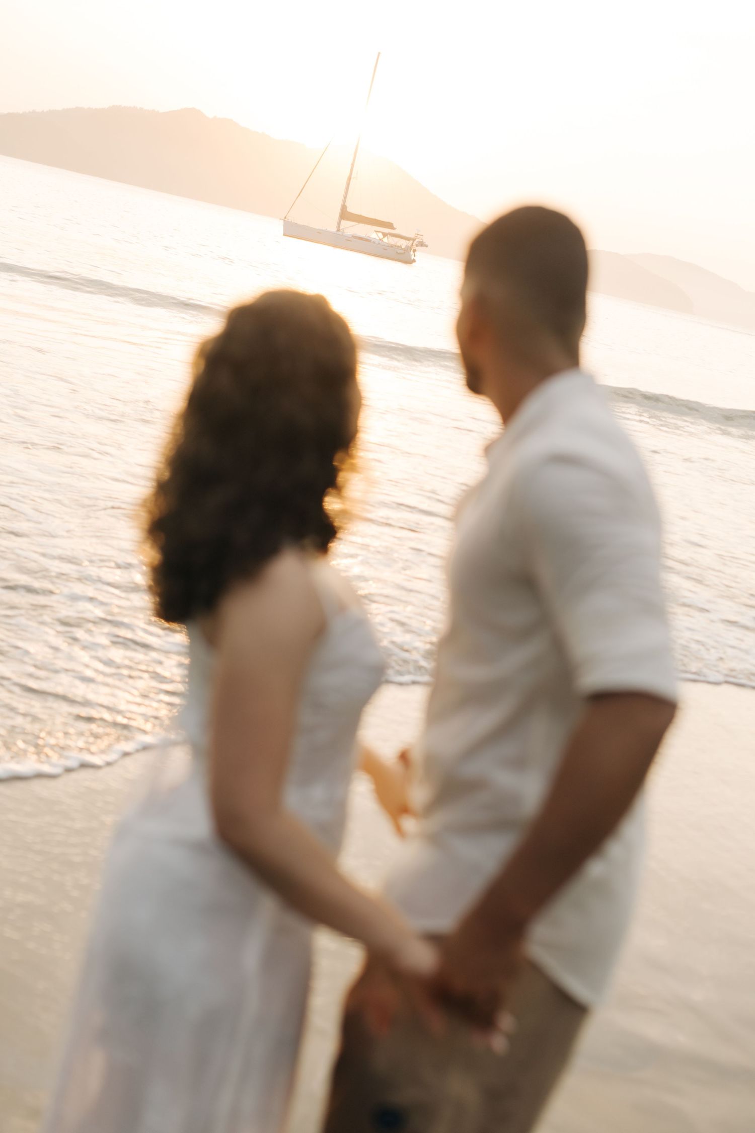 A romantic couple stands together on a beach at sunset with a sailboat in the distance.
