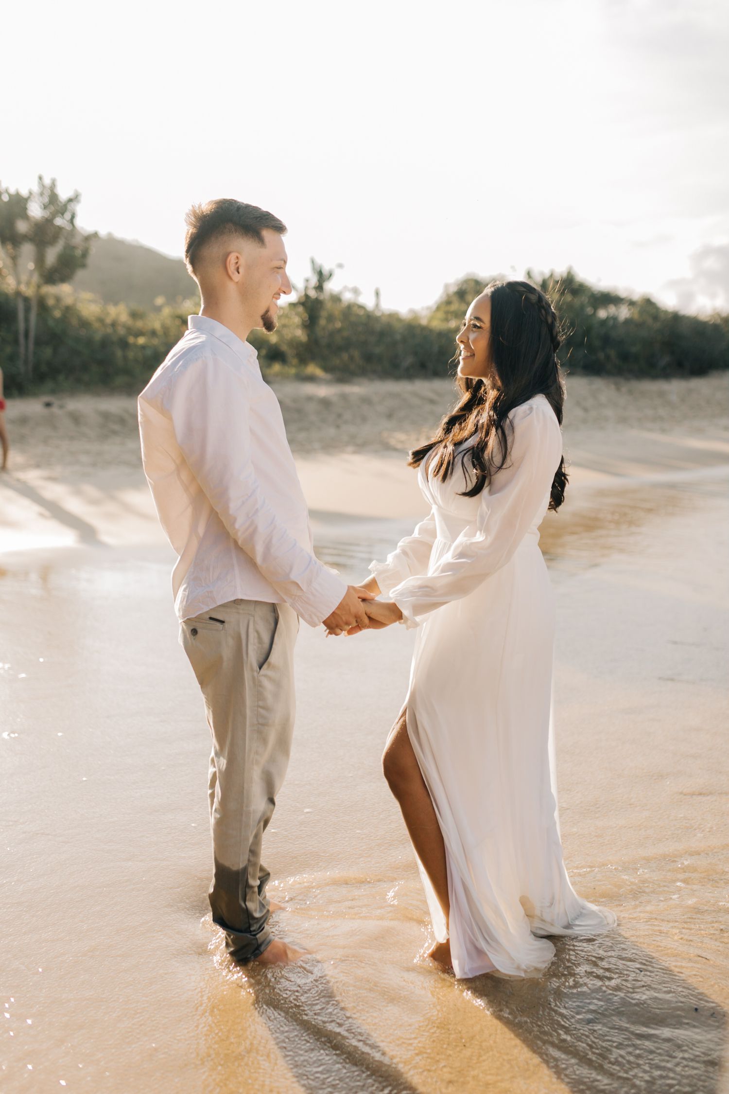 A couple holding hands stands in shallow beach water during golden hour.