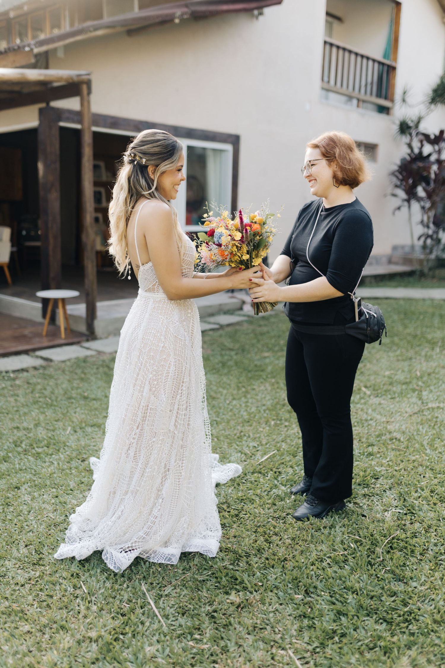 An intimate outdoor wedding scene with a bouquet handoff against a white house backdrop.