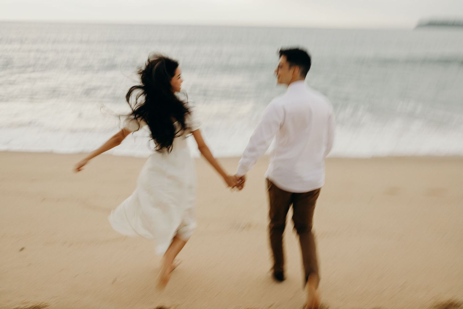 A couple runs playfully on the beach at sunset, wearing coordinated white and brown outfits.