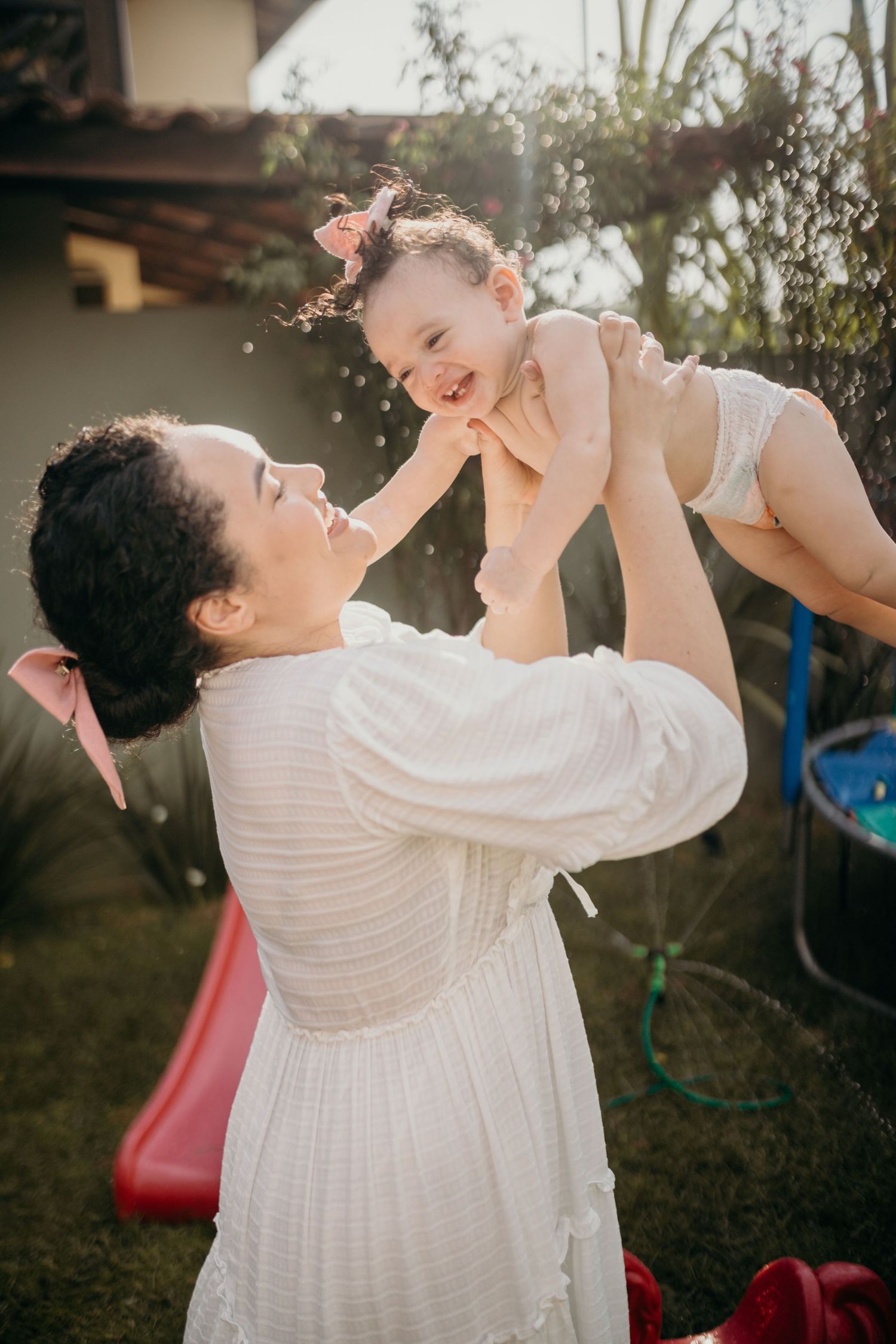 Parent in white dress lifts baby up playfully in backyard setting.