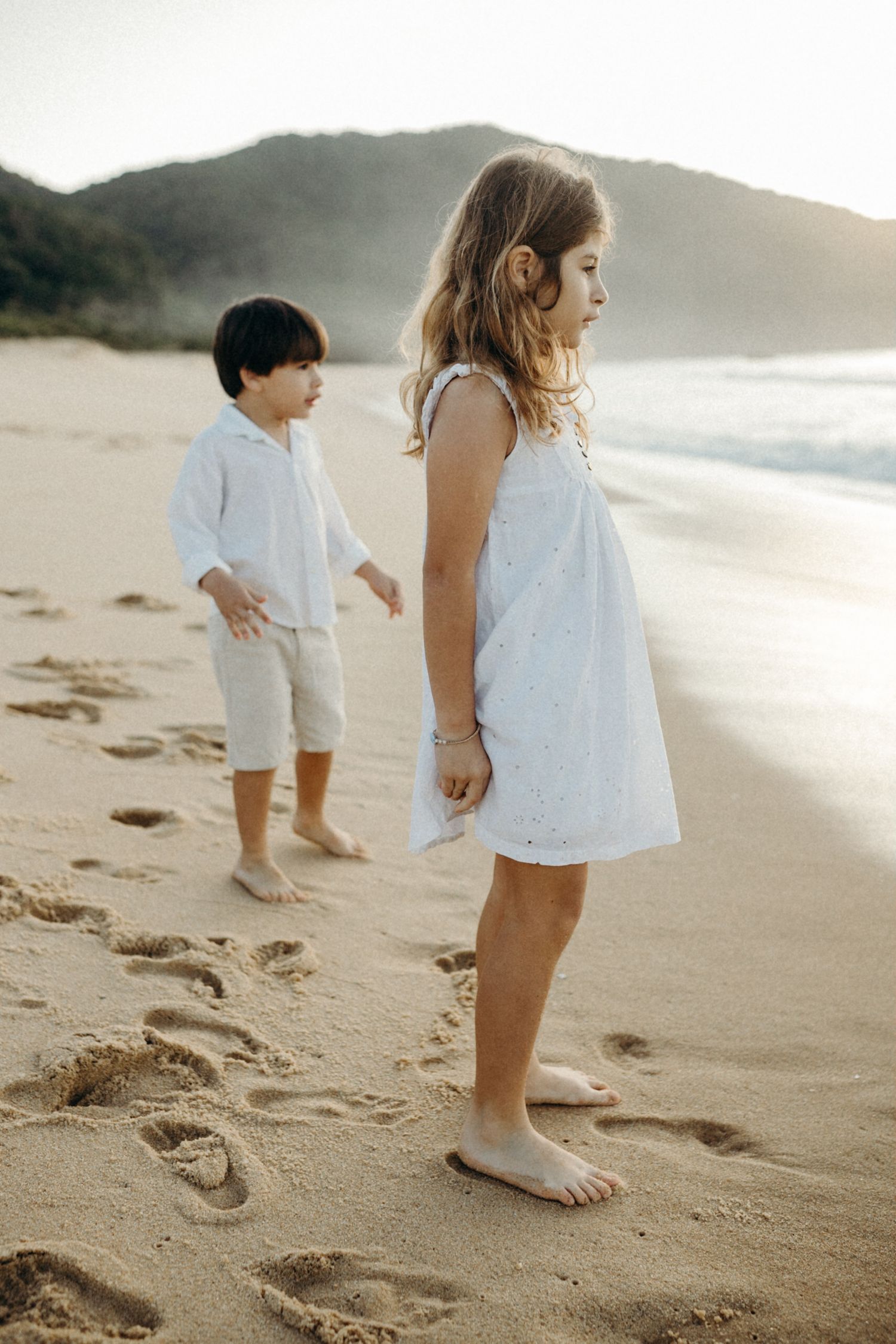 Two people in white clothing standing on a beach with mountains and mist in the background.
