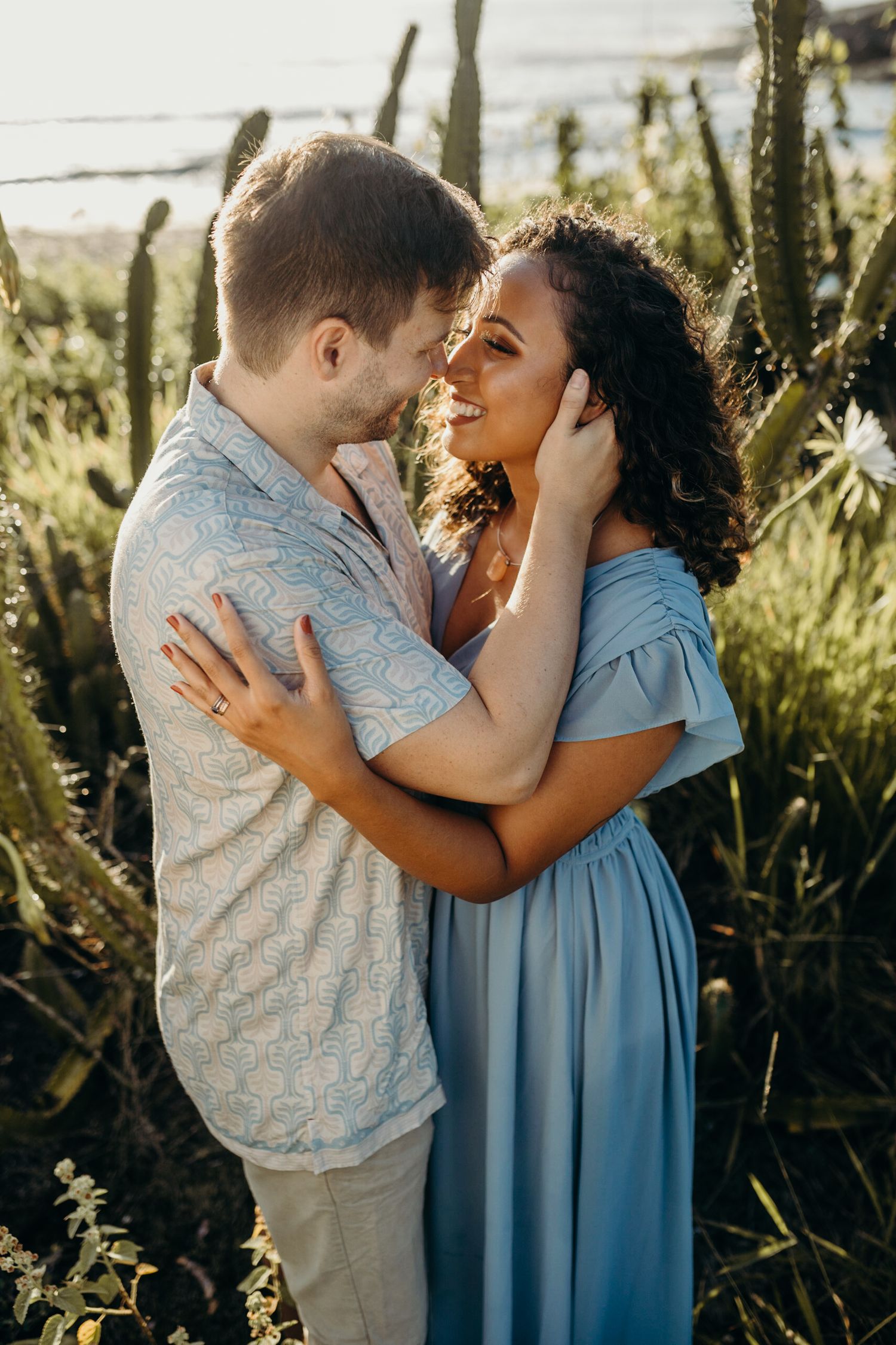 Romantic couple embraces in golden sunlight among tall grass and greenery at sunset.