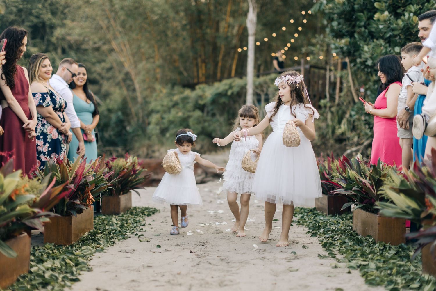 Young flower girls in white dresses walking down garden path at outdoor wedding.