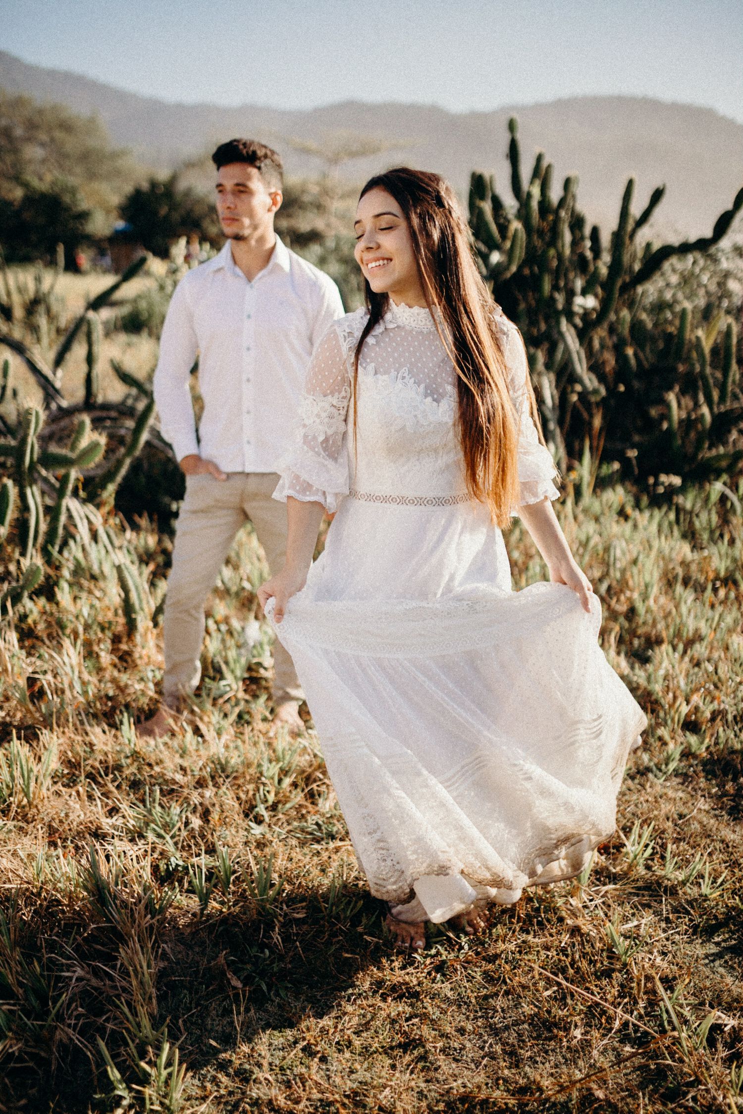 A woman in a flowing white dress walks through desert terrain while her partner stands nearby.