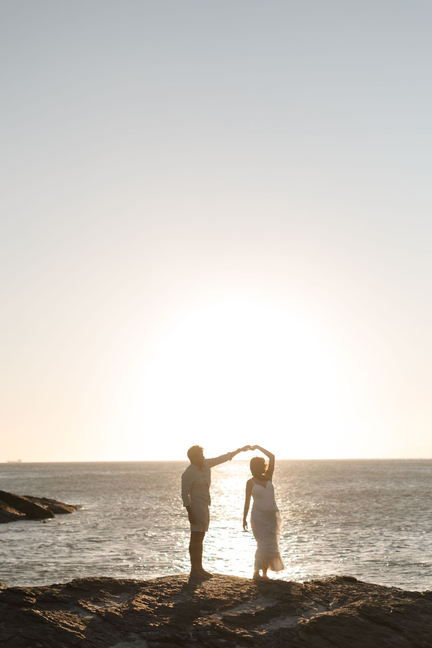 Silhouetted couple dancing on coastal rocks at sunset by the ocean.