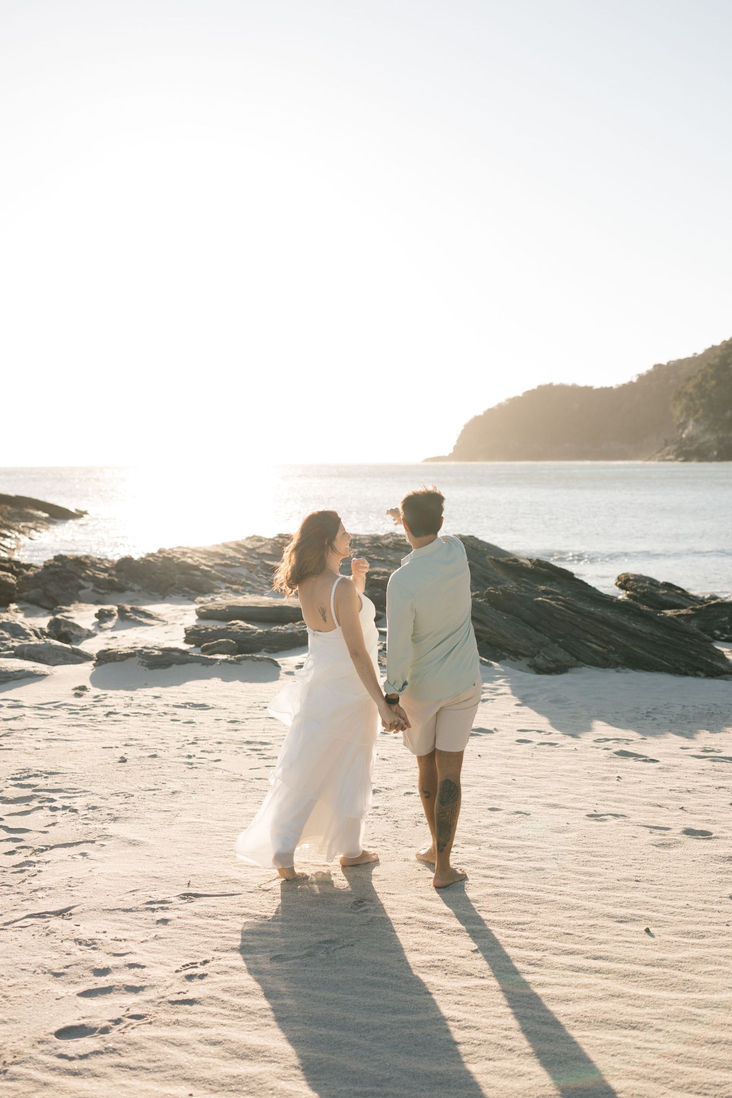 Silhouettes of a couple walking along a sandy beach at sunset with rocky cliffs in the background.