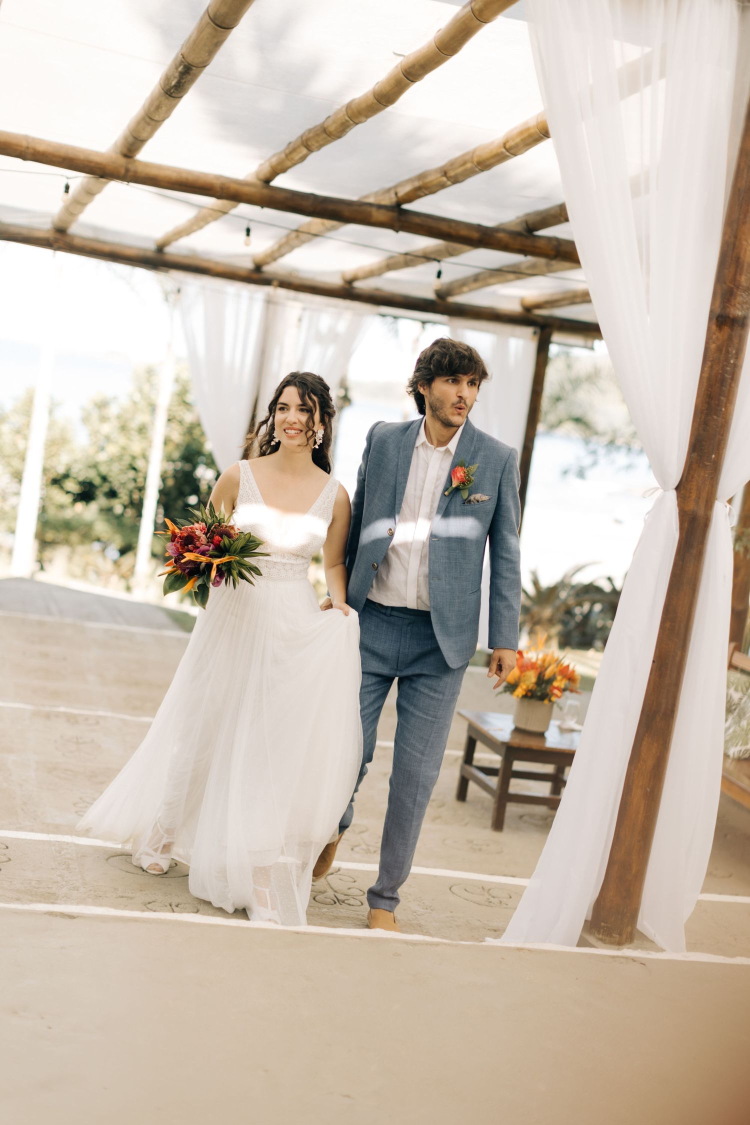 Wedding couple walking under a wooden pergola decorated with white drapes and tropical flowers.