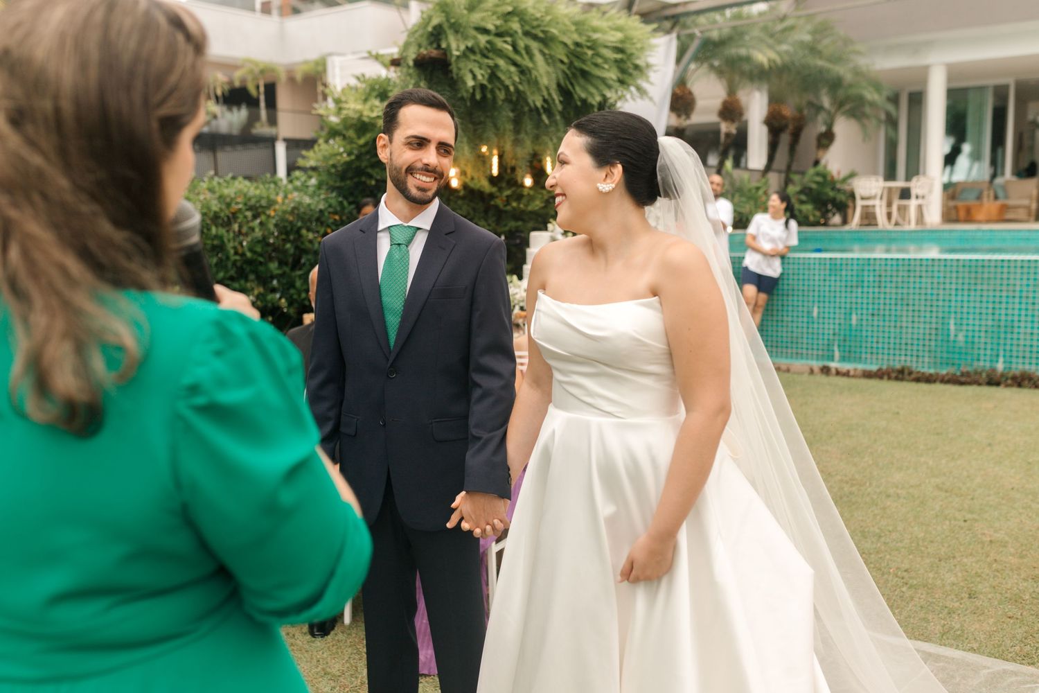 Wedding couple exchanging vows with officiant in green during outdoor ceremony.