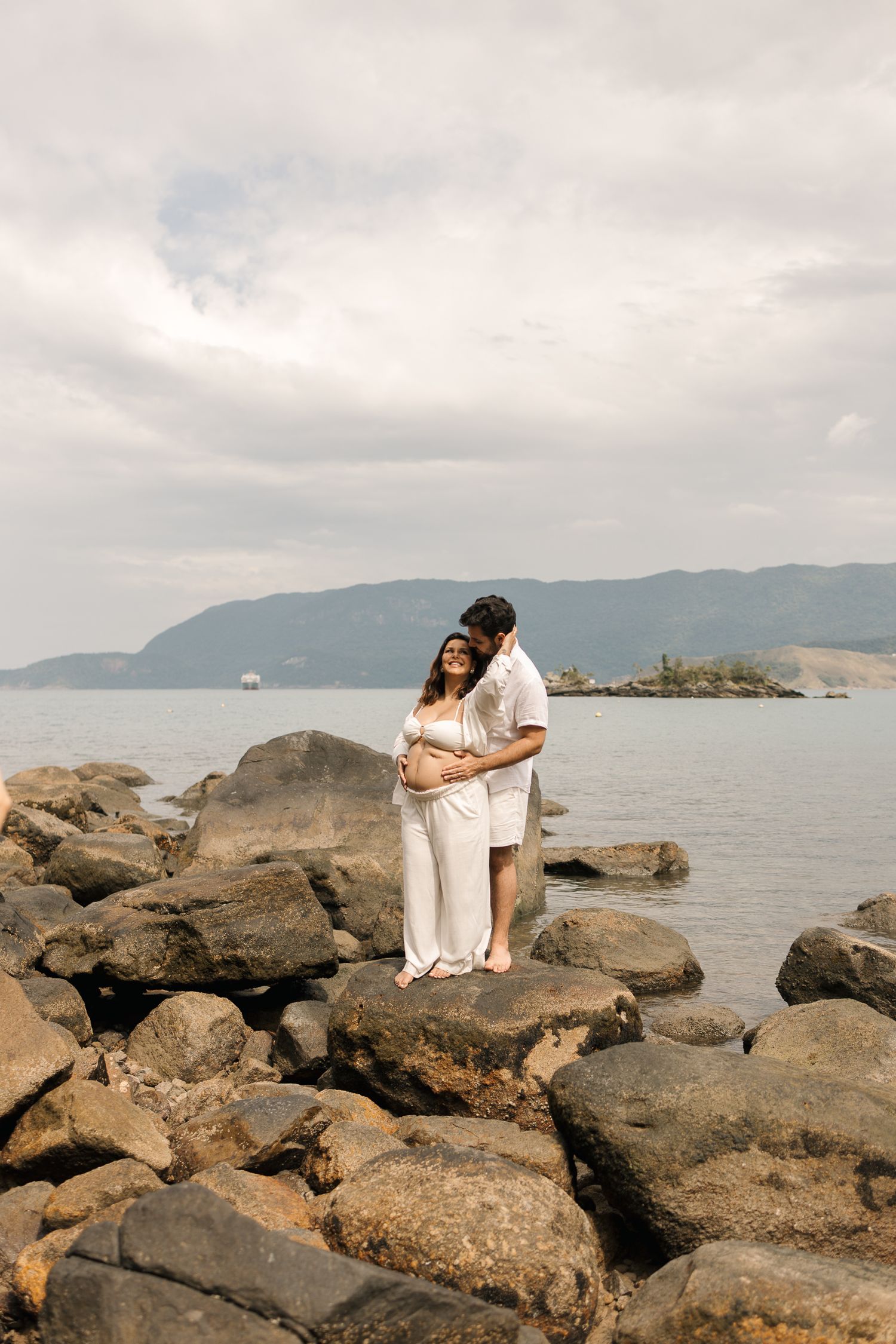 Two figures sit together on coastal rocks overlooking a misty ocean bay with mountains in the background.