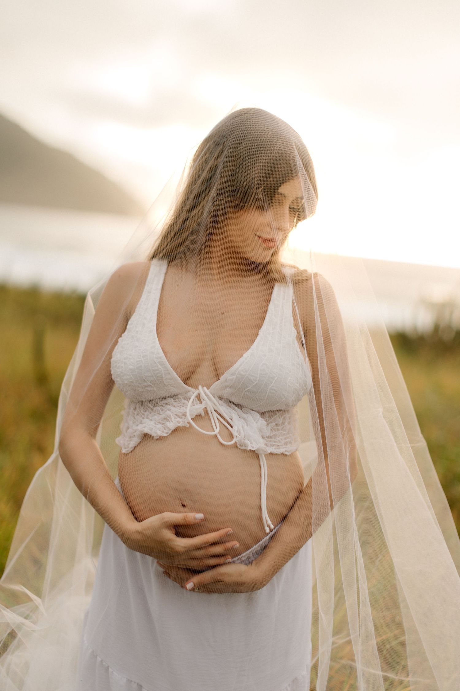 Ethereal maternity portrait of expectant mother in white lace against dreamy mountain backdrop at sunset.