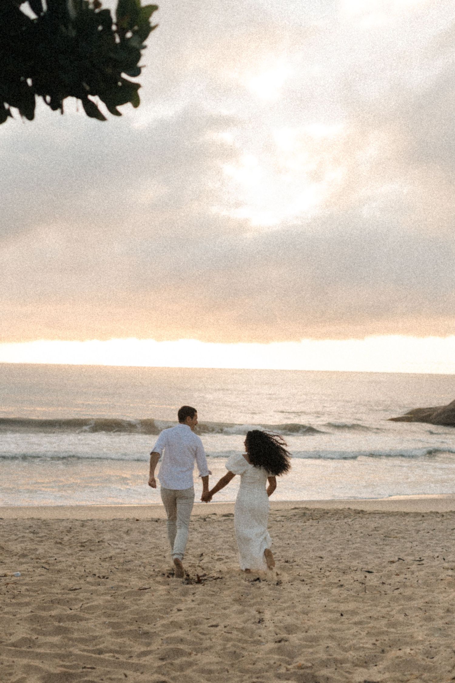 A couple runs hand in hand along a sandy beach at sunset with ocean waves in the background.