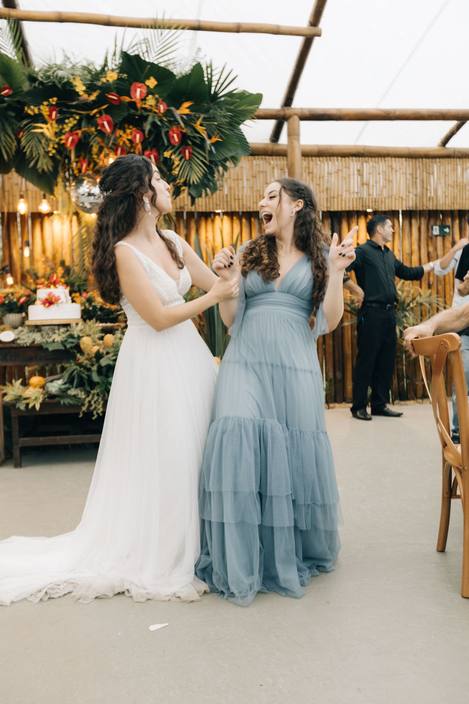 A wedding celebration shows a bride in white and bridesmaid in light blue sharing a joyful moment together.