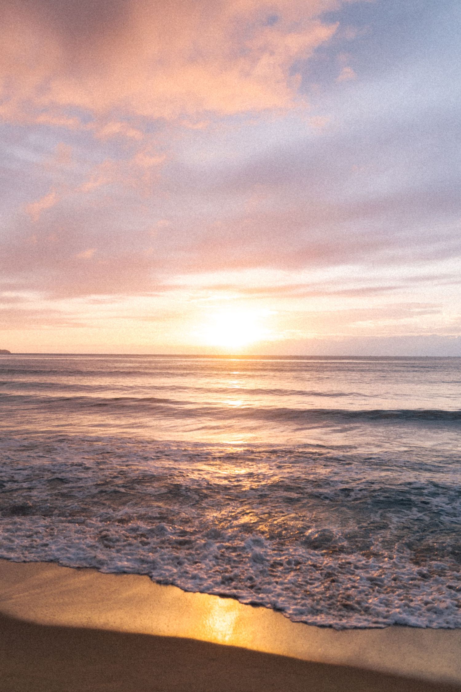 Pôr do sol dourado sobre as ondas do mar batendo na praia com nuvens rosa e roxas no céu dramático.