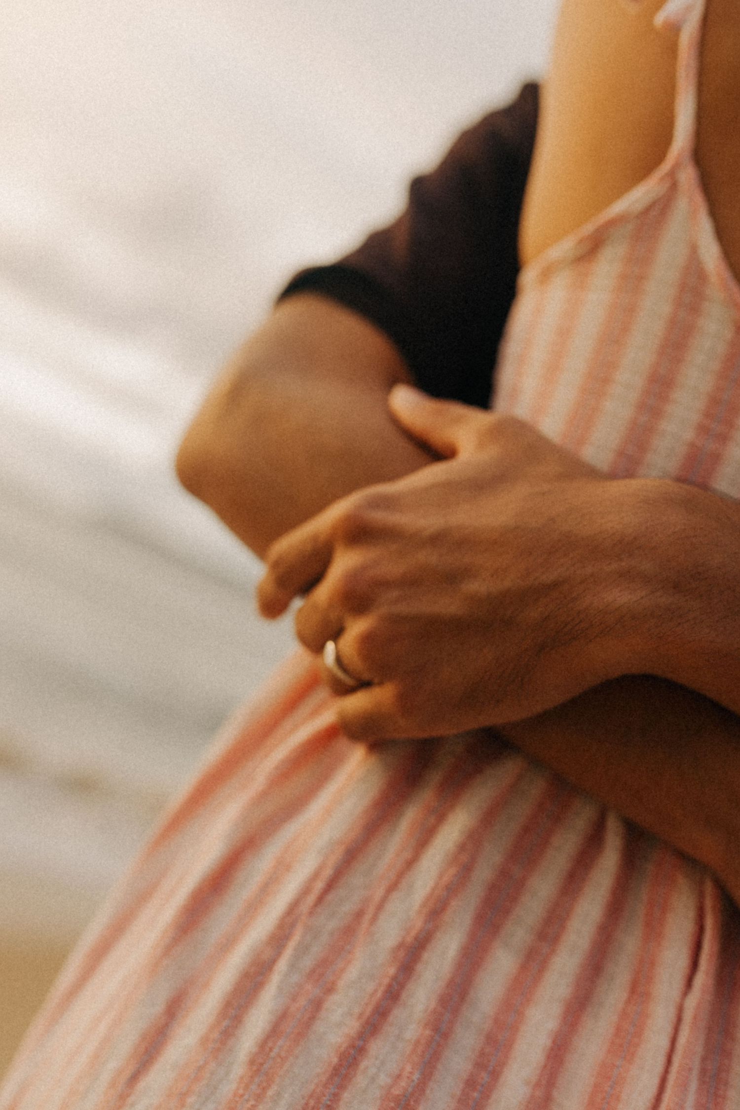 Close de mãos abraçando uma pessoa usando um vestido listrado rosa e branco, com um anel de ouro visível.