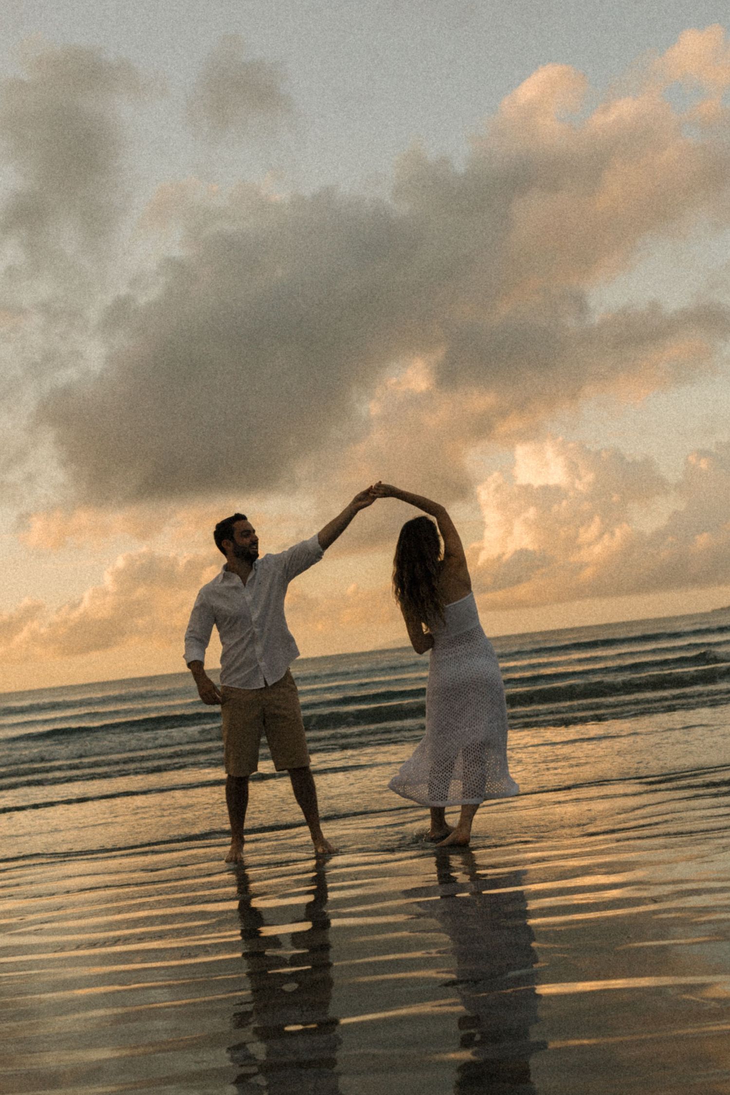 Two people dance joyfully in the reflecting waters of a beach at sunset.