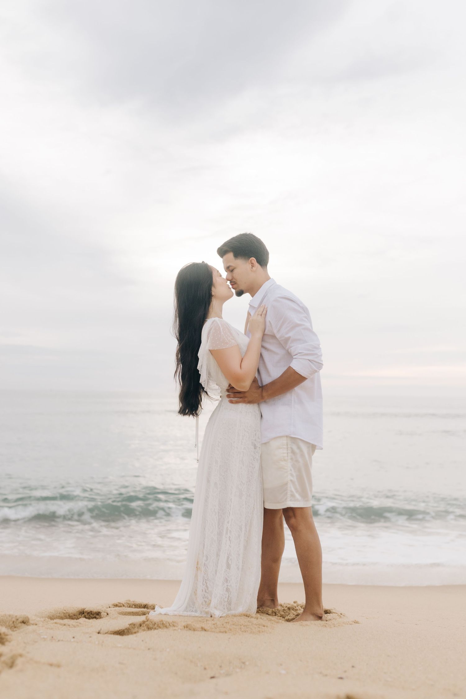 Couple in white clothing embracing on beach with waves and cloudy sky background.