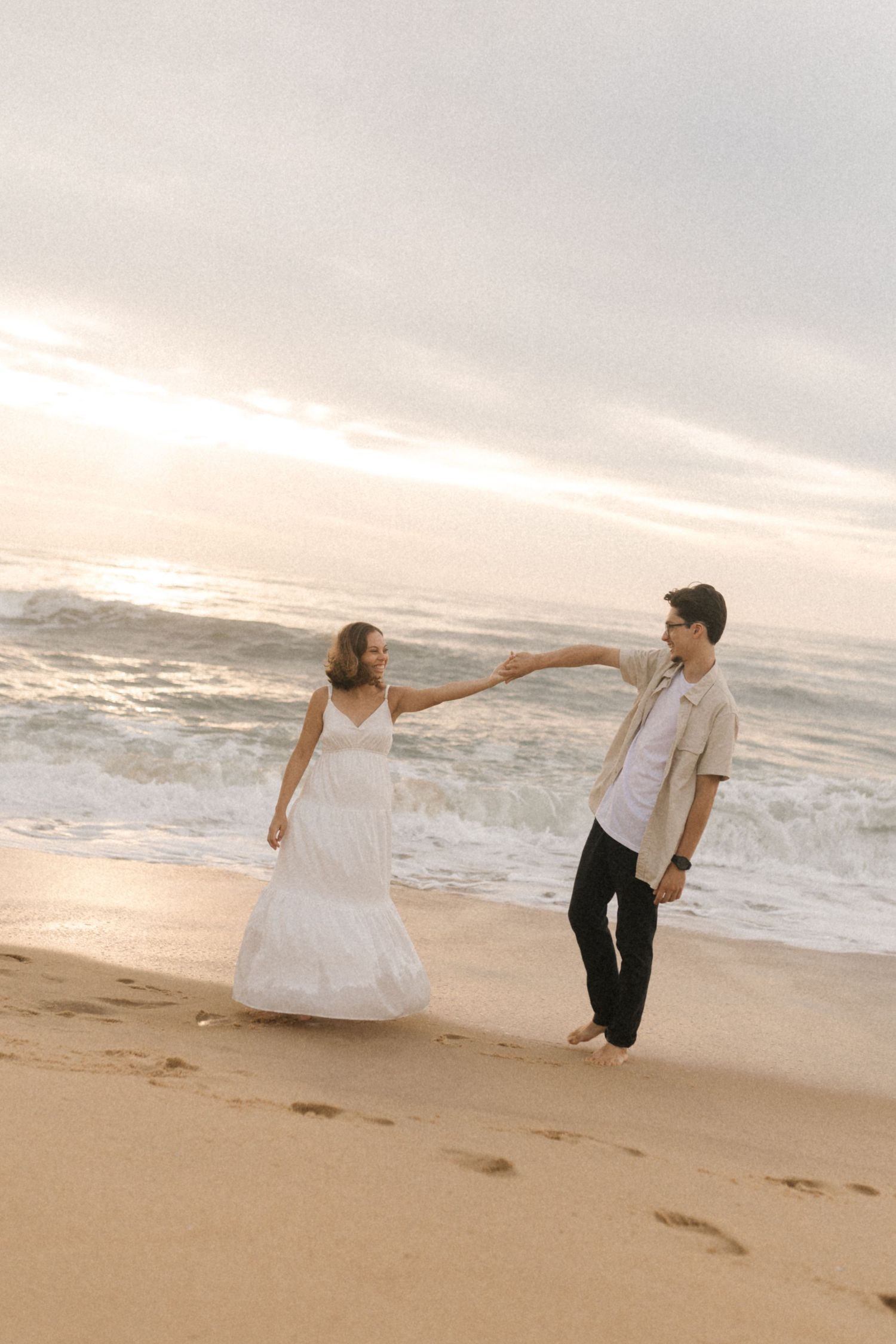 Playful couple dancing and twirling on the beach with waves crashing in the background at sunset.