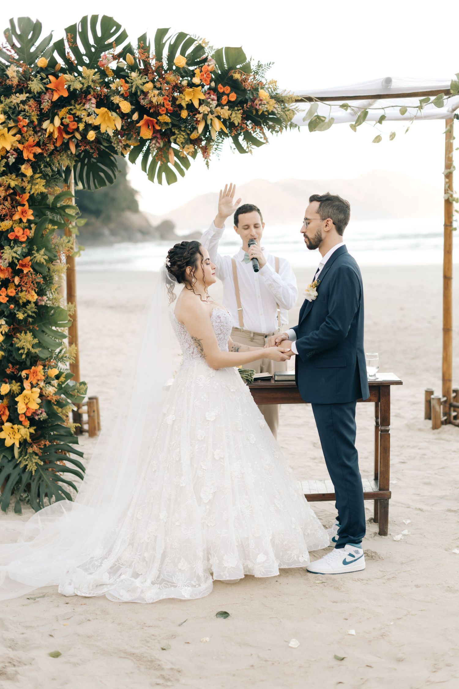 Wedding ceremony under tropical floral arch with monstera leaves and orange blooms.