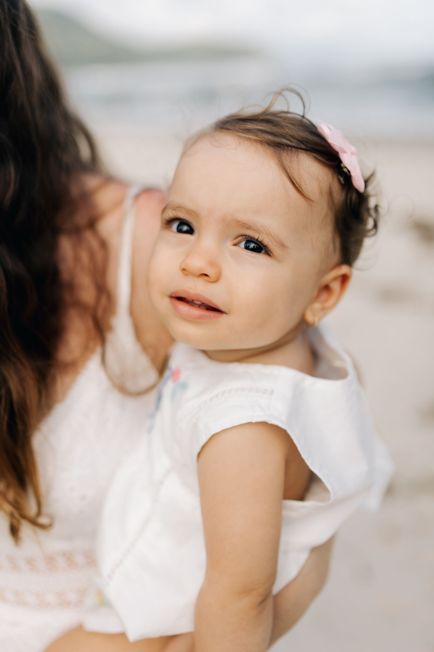 Close-up of baby in white dress with tiny pink hair bow looking up sweetly.