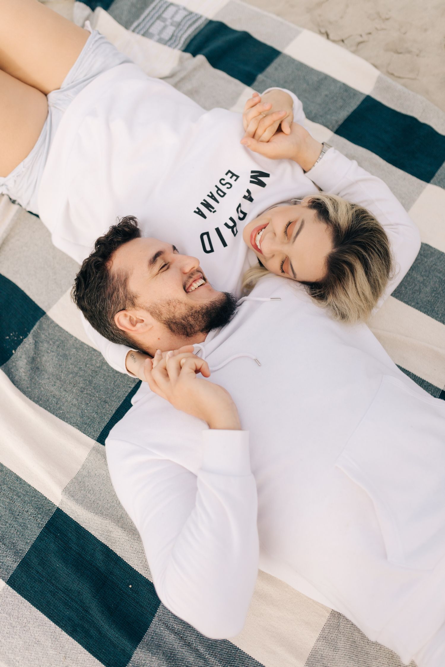 Couple in white clothing sharing joyful moments while lying on a striped blanket.
