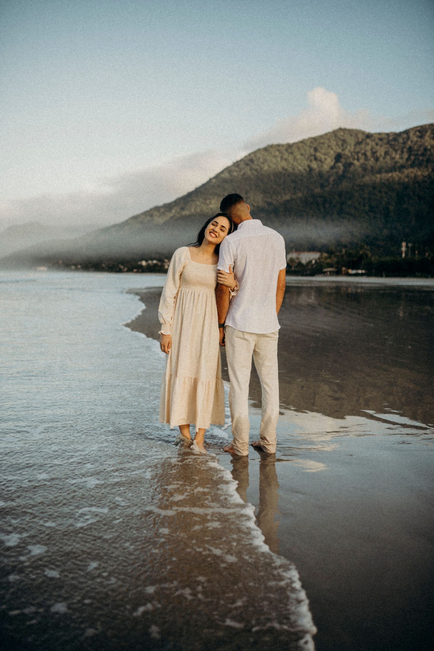 Couple in cream colored outfits stands together on misty beach with mountain backdrop.