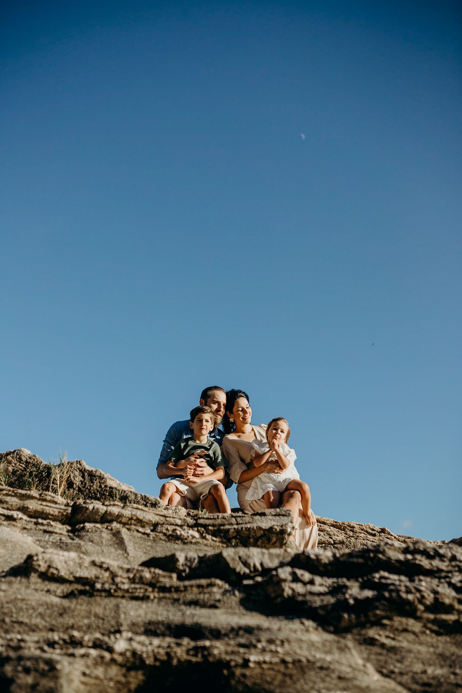 Group relaxing on rocky cliff overlooking blue ocean waters under clear sky.