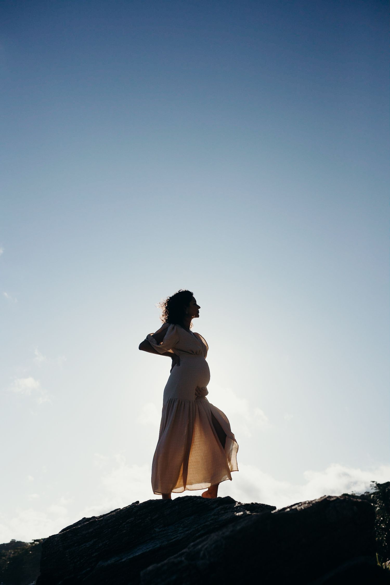 Backlit silhouette on rocky cliff wearing flowing dress in breeze.