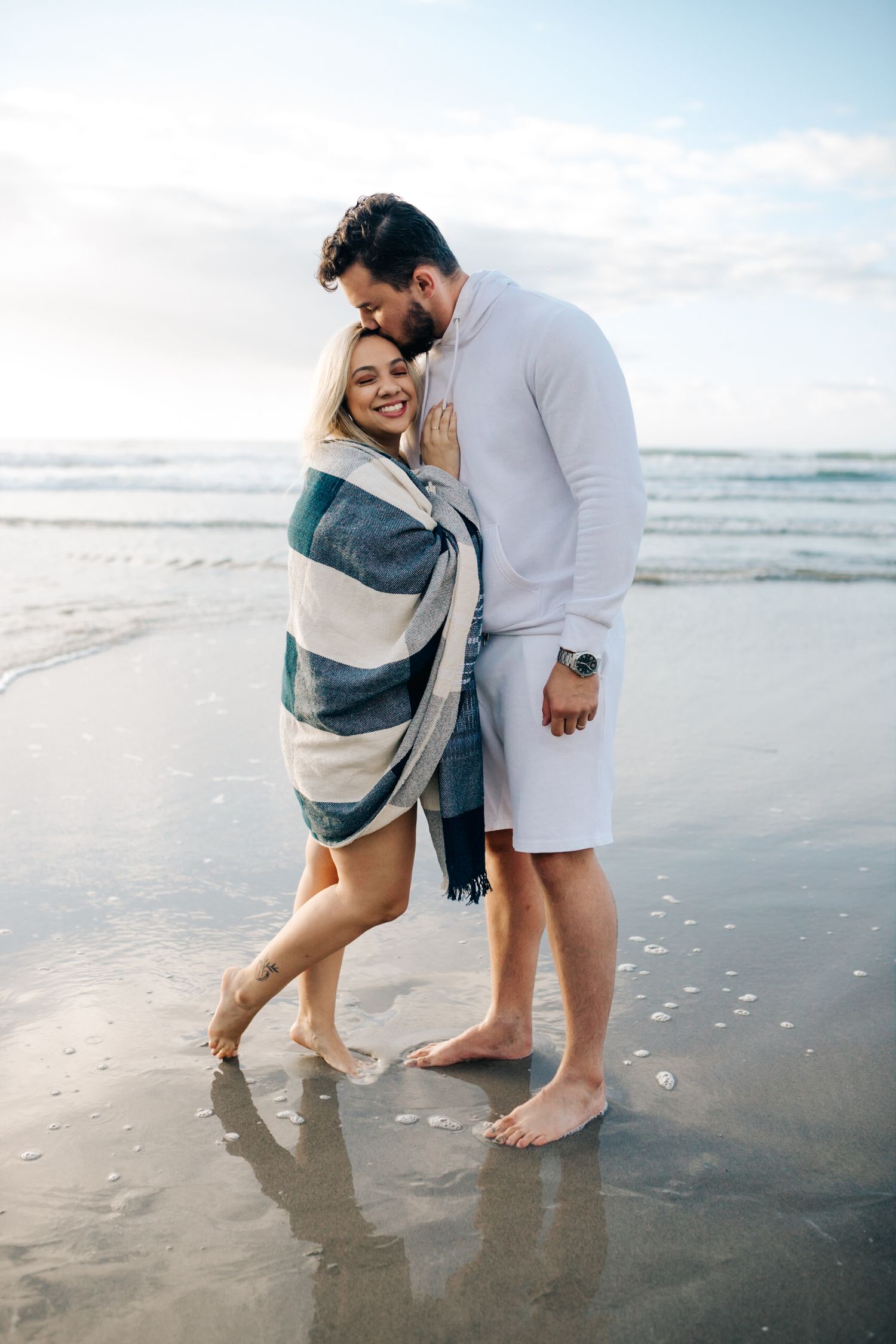 Couple sharing an intimate moment while wrapped in a striped blanket on a misty beach at sunset.
