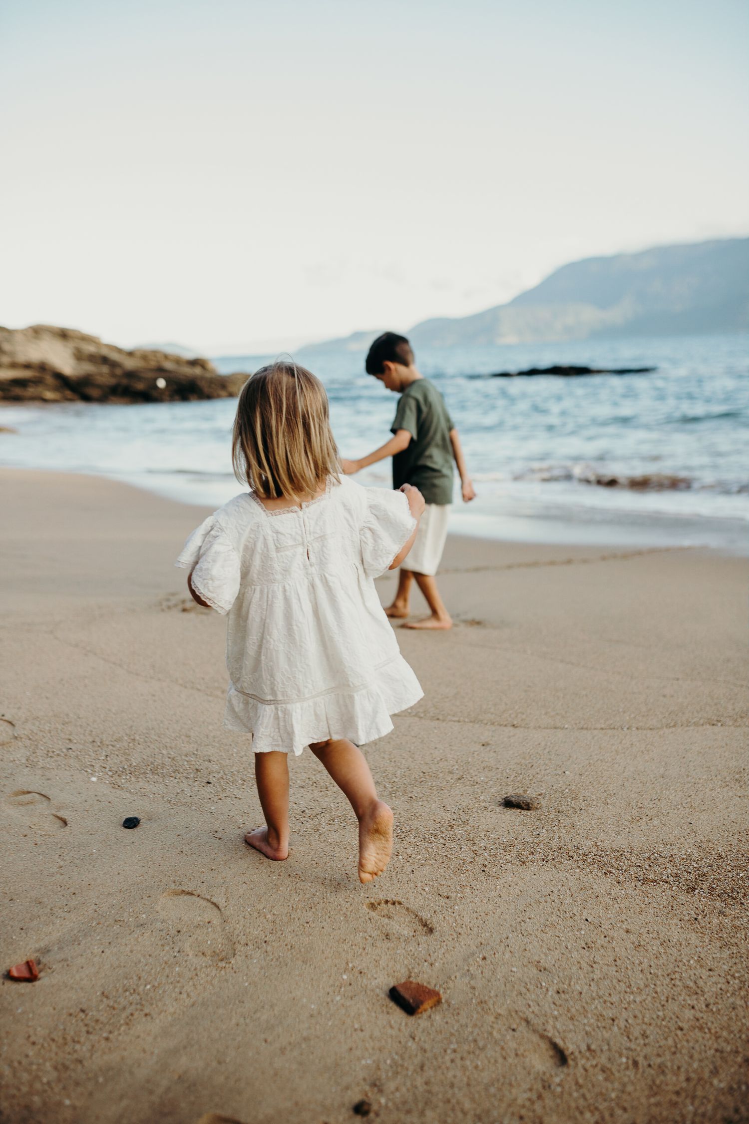 A small child in a white dress runs playfully on a sandy beach.