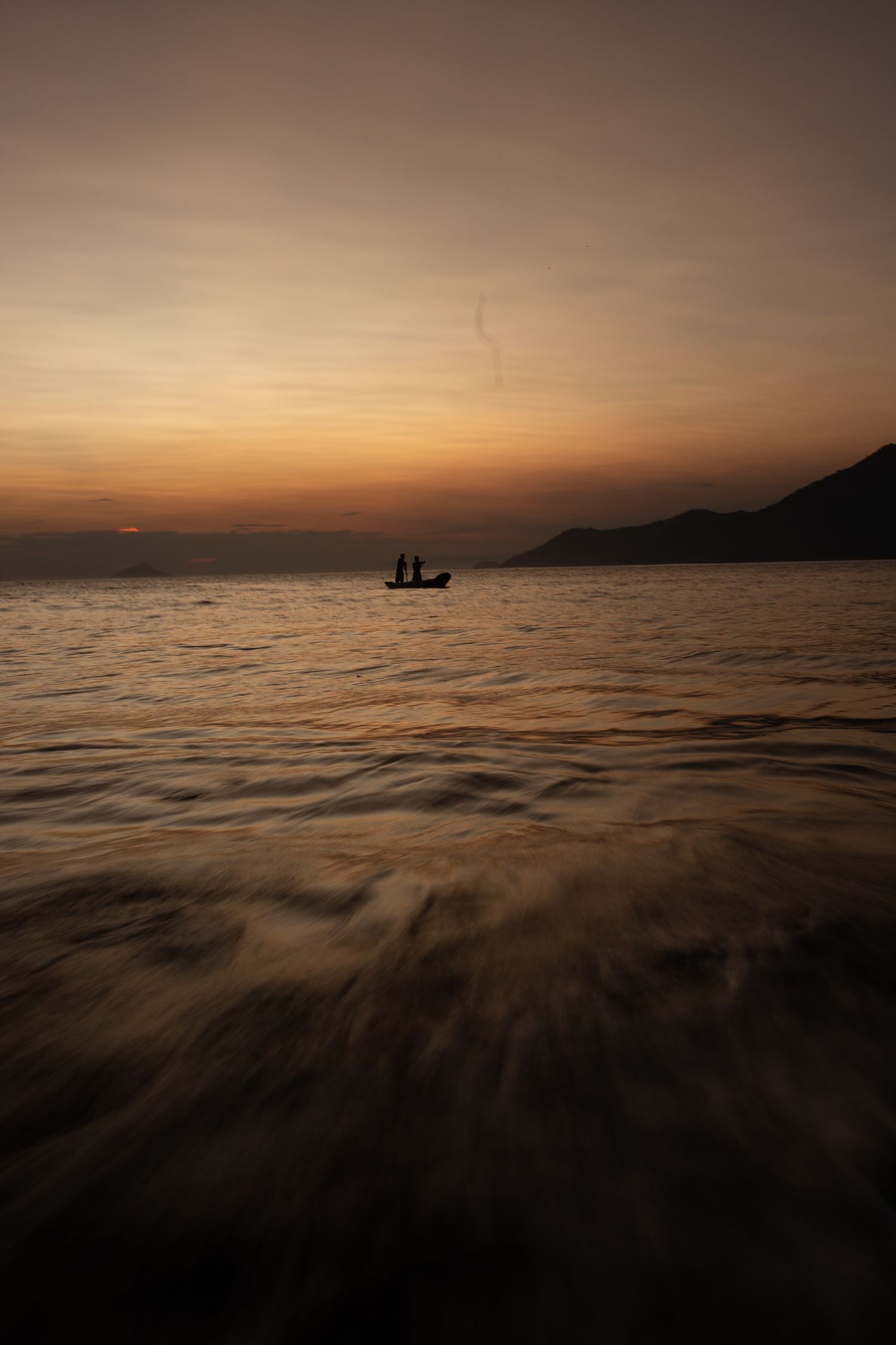 Small boat silhouette on calm waters during golden sunset with mountains in the background.