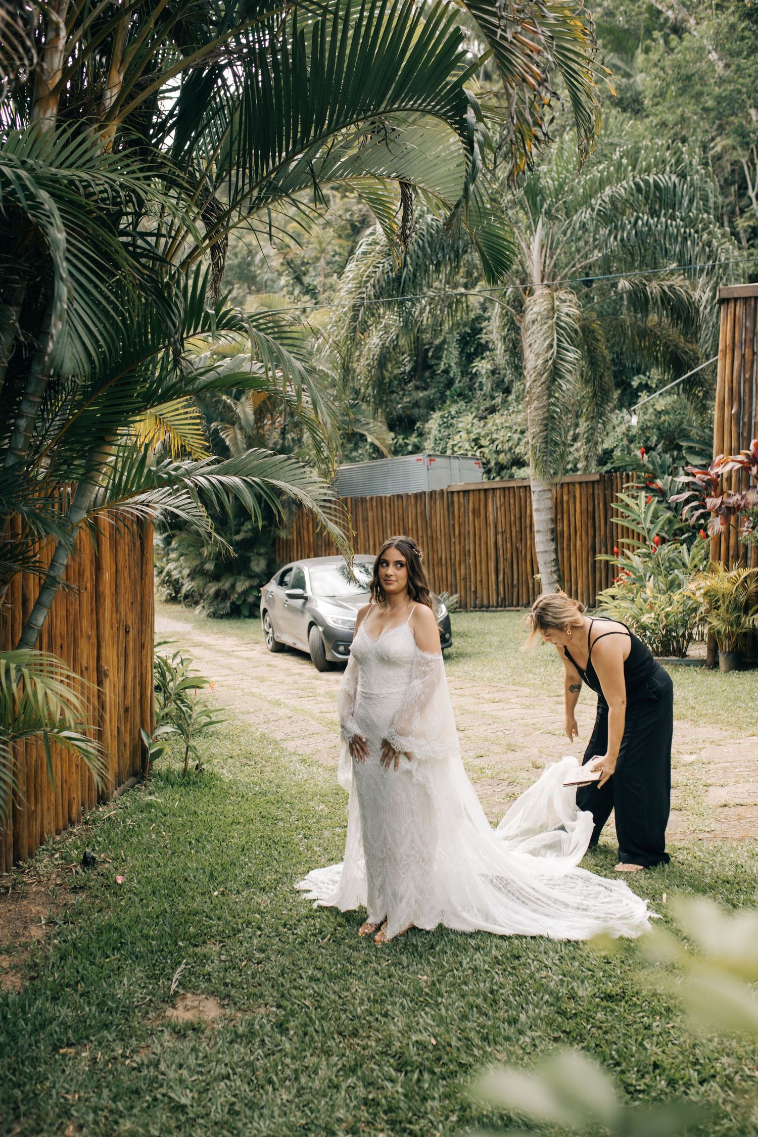 A wedding dress being arranged in a lush tropical garden setting with palm trees and bamboo fencing.