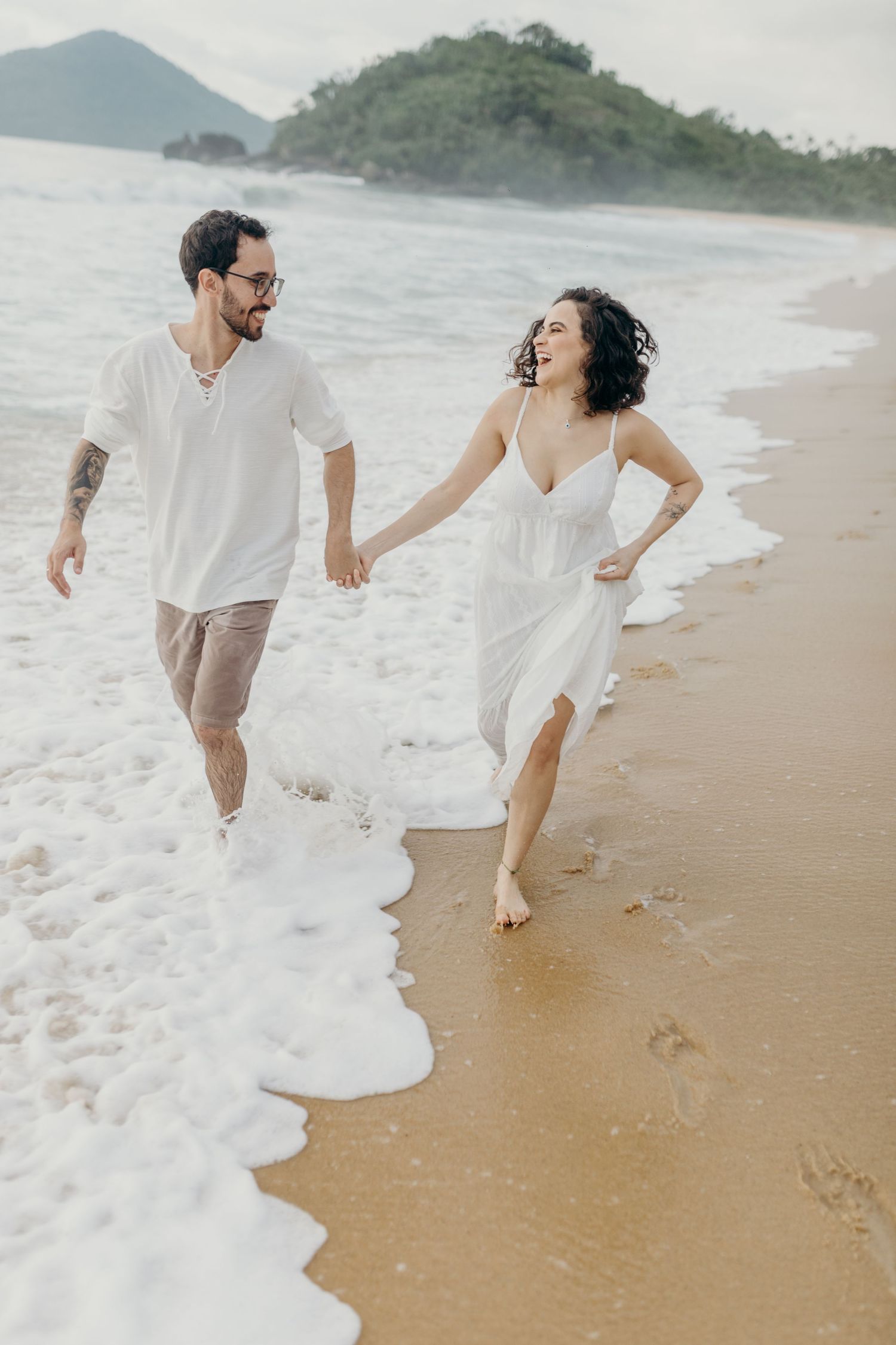 A couple runs playfully along the shoreline with mountains and greenery in the background.