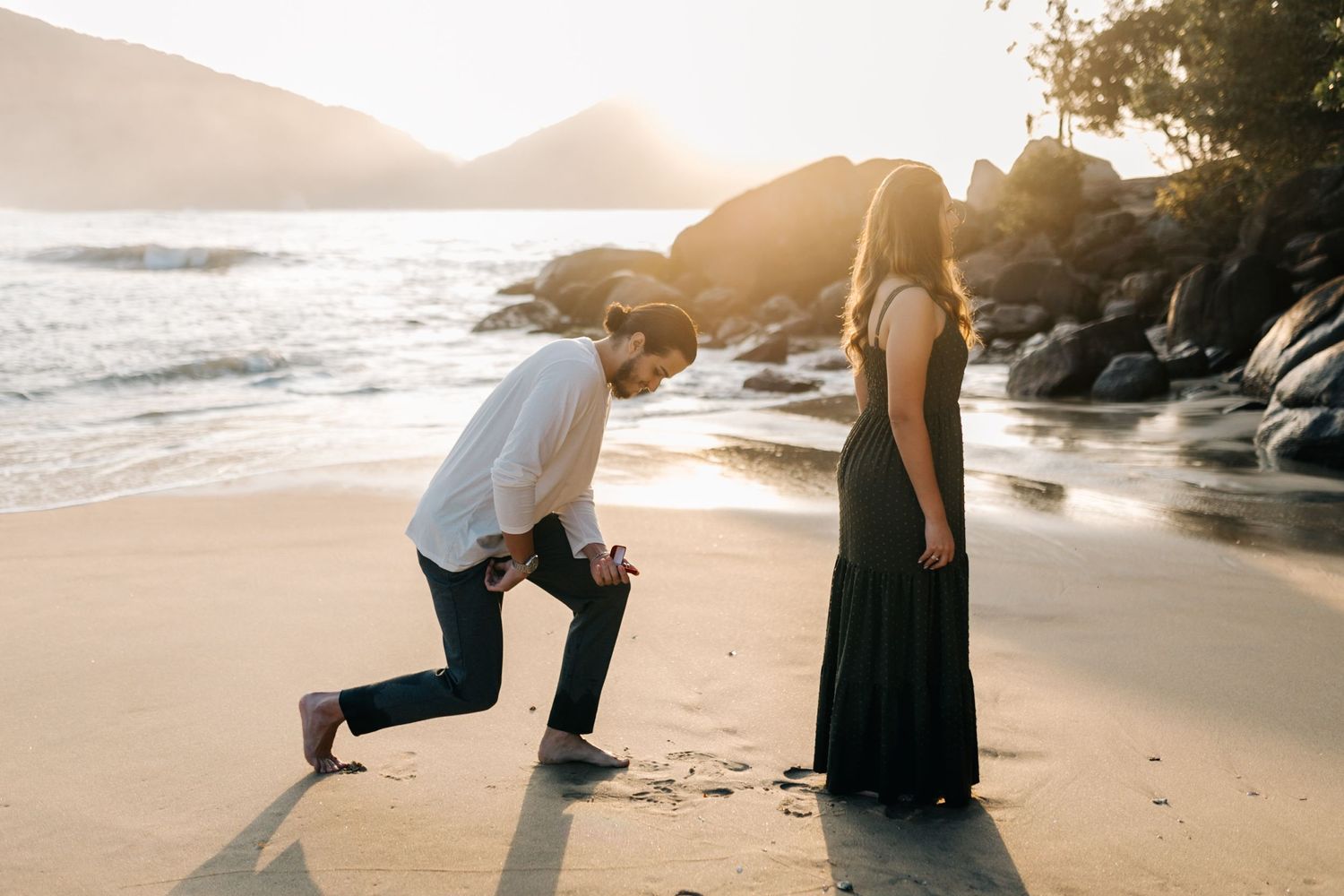 Proposal moment captured on a scenic beach at sunset with rocky coastline in the background.