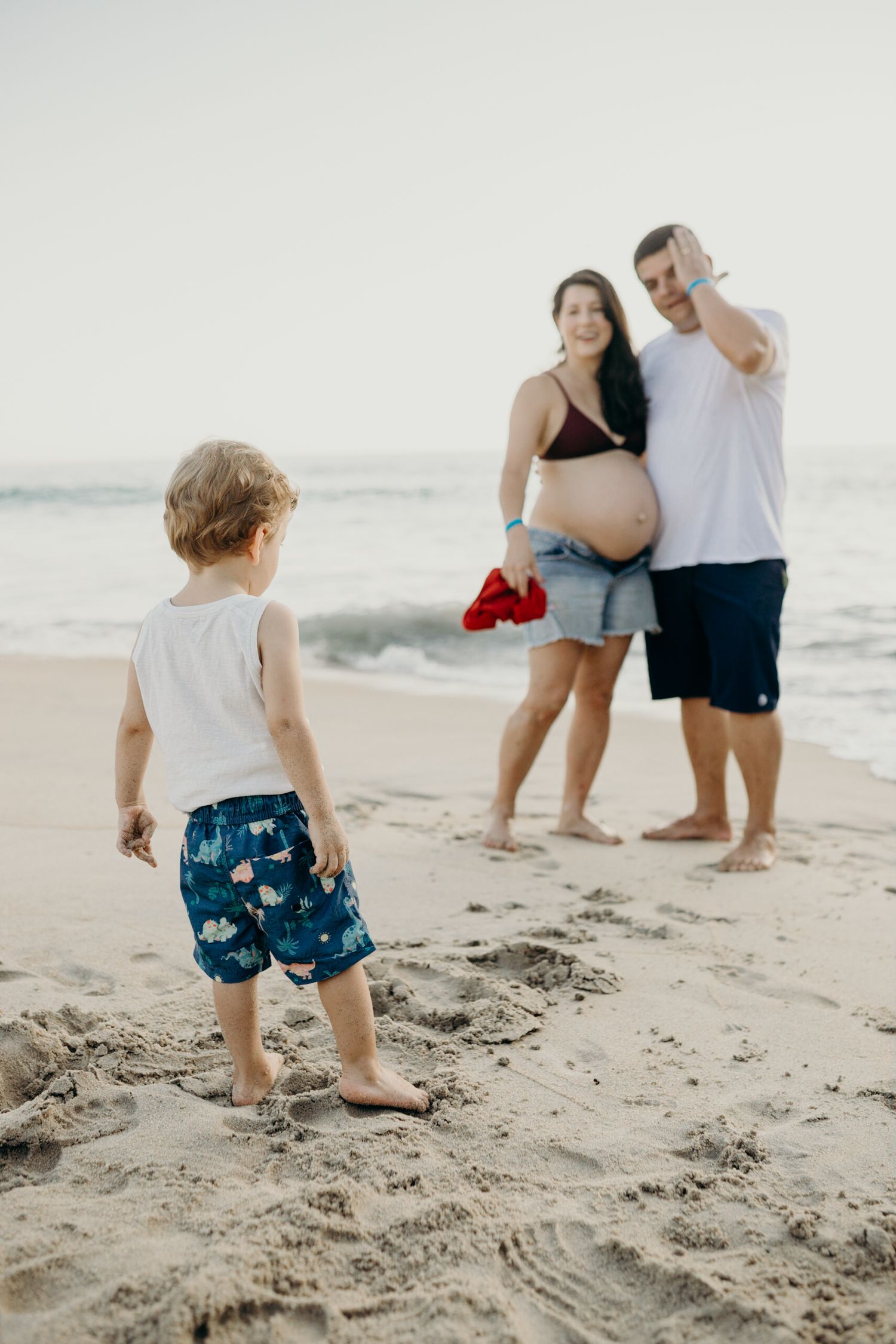 A young child in blue swim shorts plays in the sand while adults watch from behind.